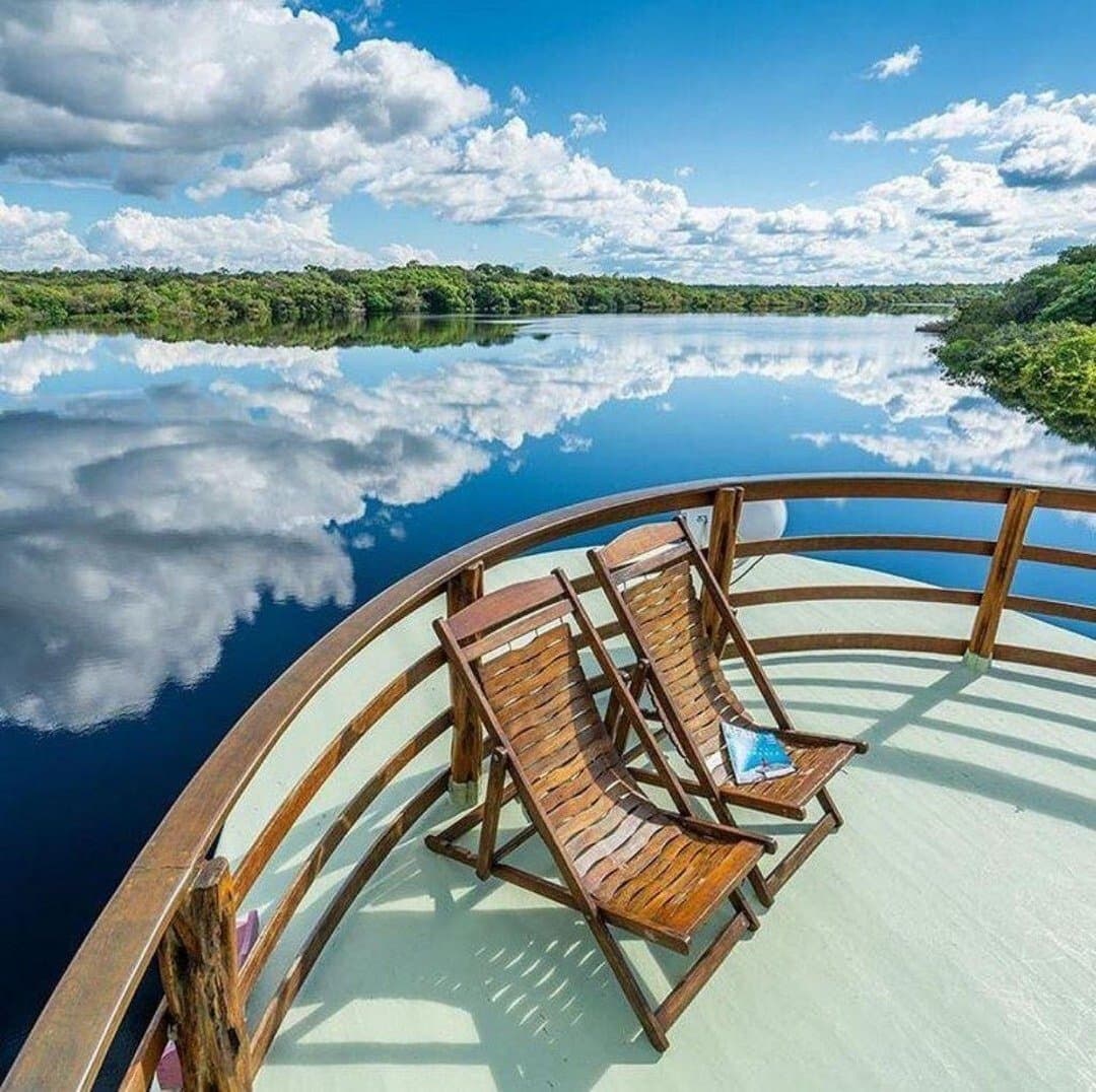 Your seat is ready. The river will do the rest. Two wooden deck chairs on the bow of an Amazon jungle cruise Brazil riverboat, facing a glassy black-water river perfectly mirroring a blue sky filled with white clouds and dense forest on both banks