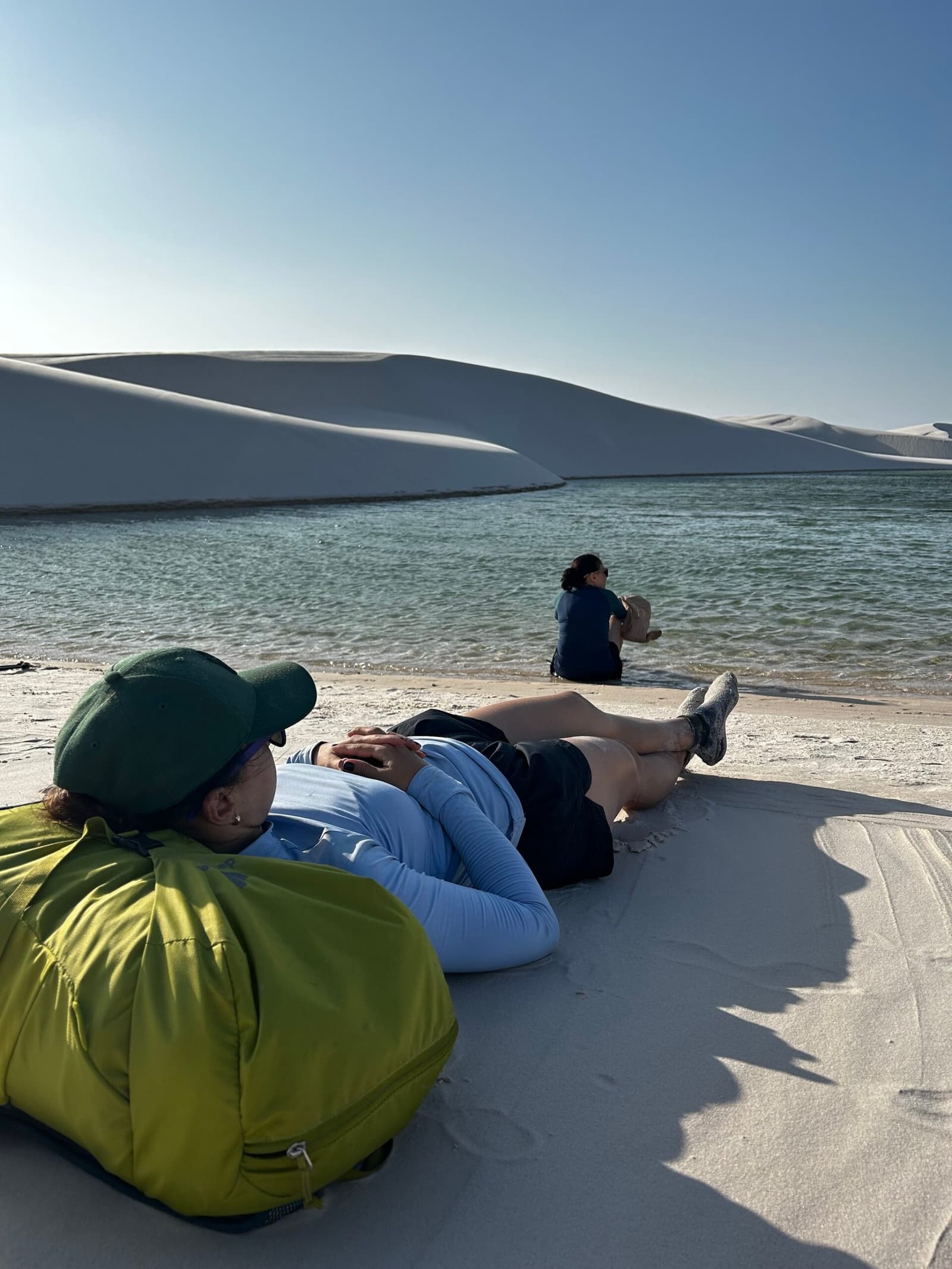 Some places earn a long horizontal pause. A hiker lying back on white sand dunes during a Lençóis Maranhenses hike, resting beside a clear freshwater lagoon while a second person sits at the water's edge, with sweeping white dunes rising behind