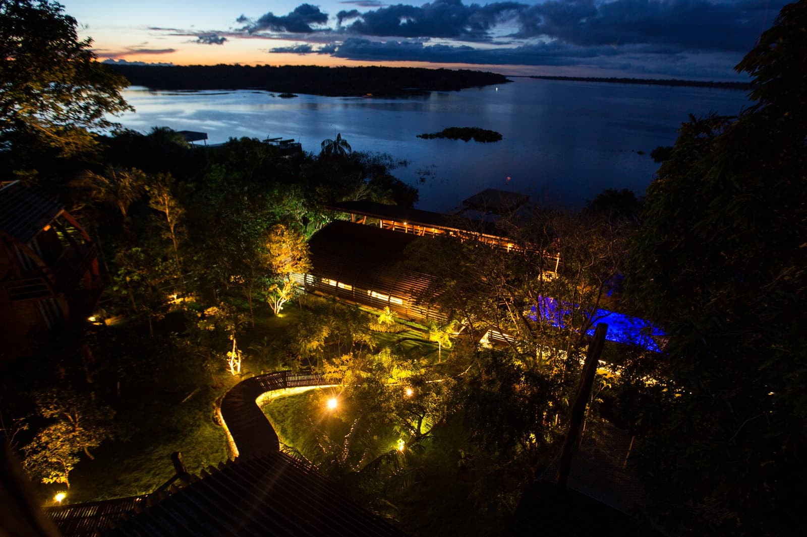The hour when the river turns gold and the lodge lights up like a lantern in the forest. Aerial twilight view of Mirante do Gavião amazon lodge with warm garden lighting, a winding boardwalk, illuminated pool, and the broad Rio Negro stretching to the horizon under a deep blue dusk sky