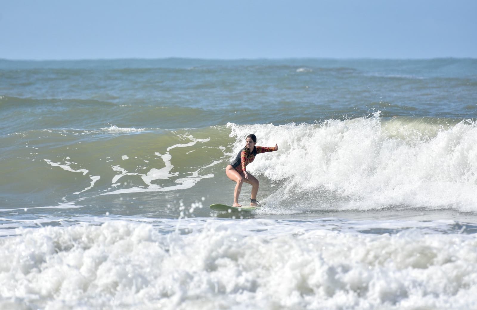 Locked in, arms out, reading the wave like she wrote it. Photo: Picasa A woman in a patterned long-sleeve rash guard rides a breaking wave during surf lessons in Itacaré, arms outstretched for balance on a green shortboard