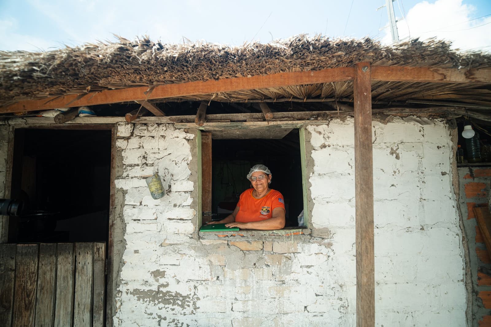 The best welcome committee needs no sign on the door. A smiling woman wearing a hairnet and orange shirt leans out of a small window in a whitewashed adobe house with a thatched palm roof, part of a local family homestay on the Lencois Maranhenses trek from Atins.