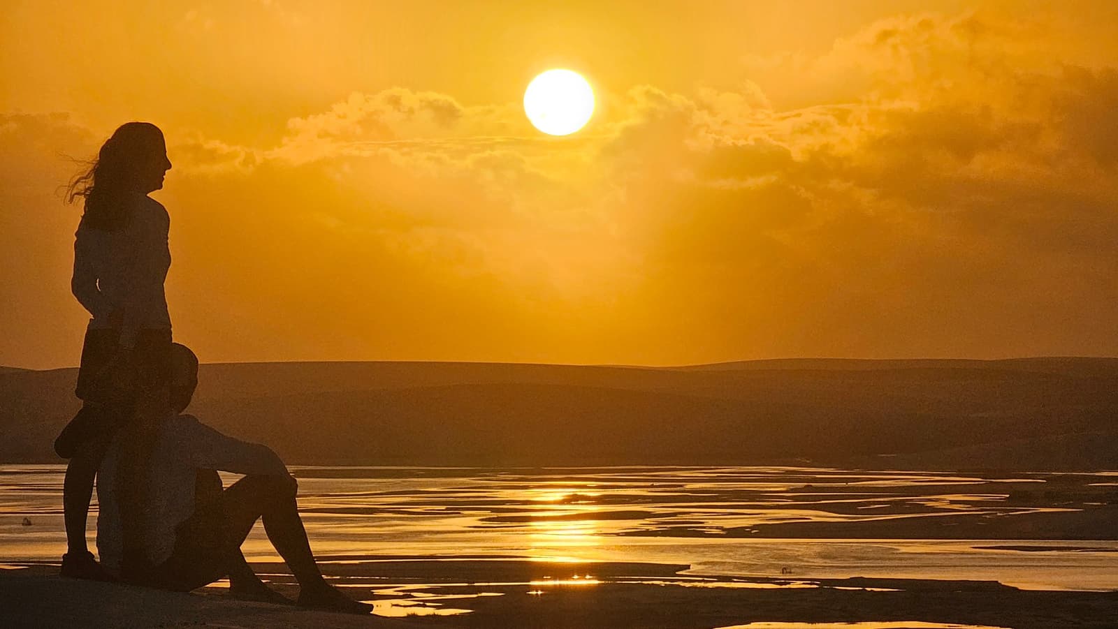 Some sunsets just make you sit down and take it all in. Two travellers silhouetted against a deep golden sunset over the flat, water-streaked dune landscape of the Lençóis Maranhenses trek, one standing and one seated