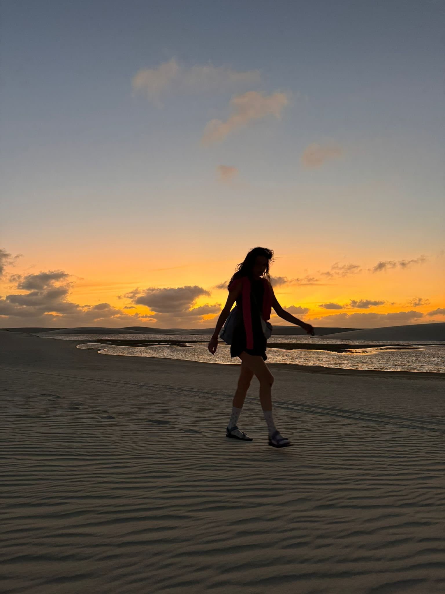 Footprints, warm wind, and nowhere else to be. Silhouette of a woman walking across rippled sand dunes during a Lençóis Maranhenses hike, with a glowing orange sunset and a shallow lagoon reflecting the sky behind her