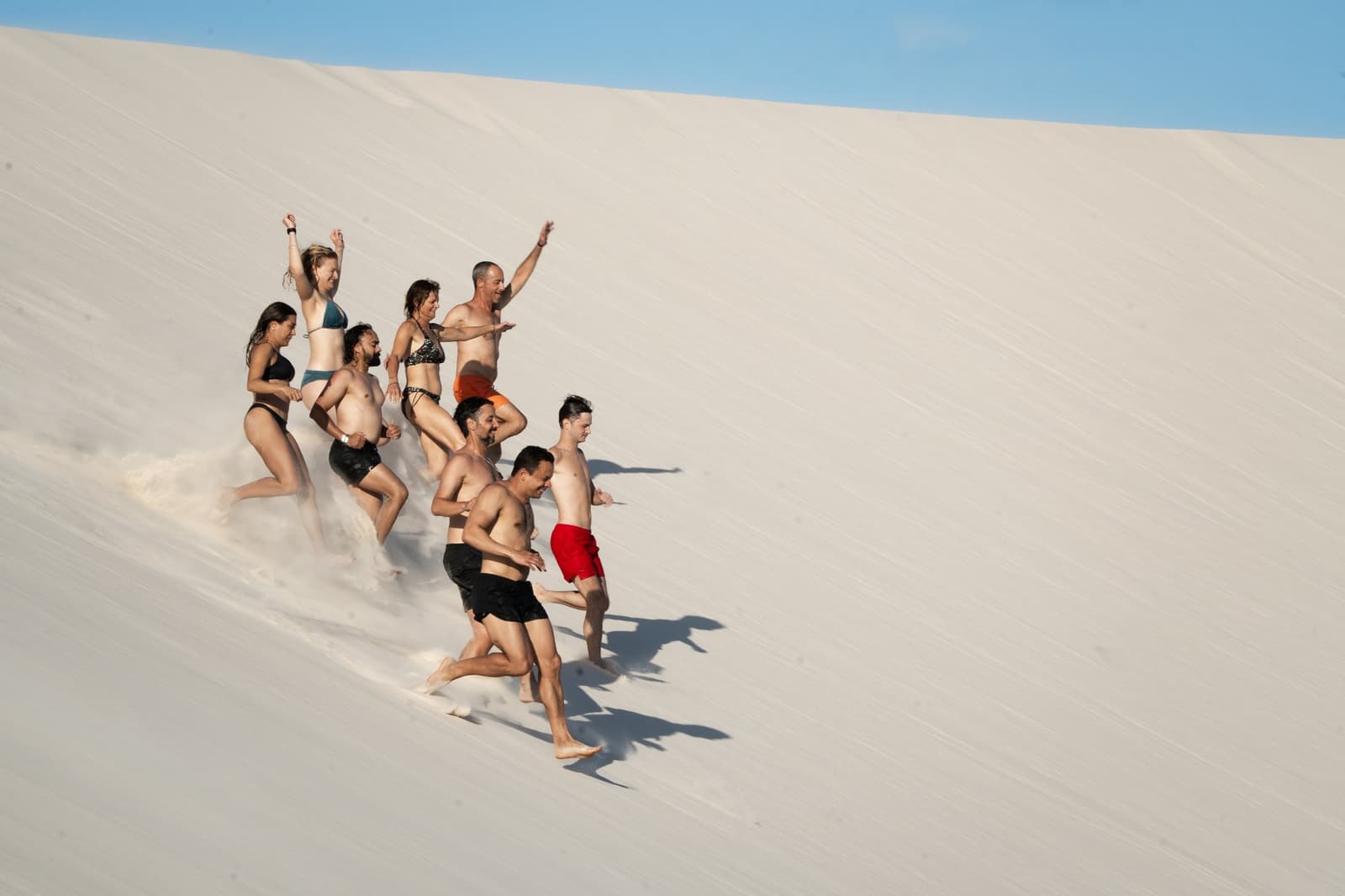 Racing down a dune is basically mandatory at this point. A group of eight travellers in swimwear running and laughing down a steep white sand dune on the Lençóis Maranhenses trek from Atins, arms raised and sand flying beneath their feet under a clear blue sky.