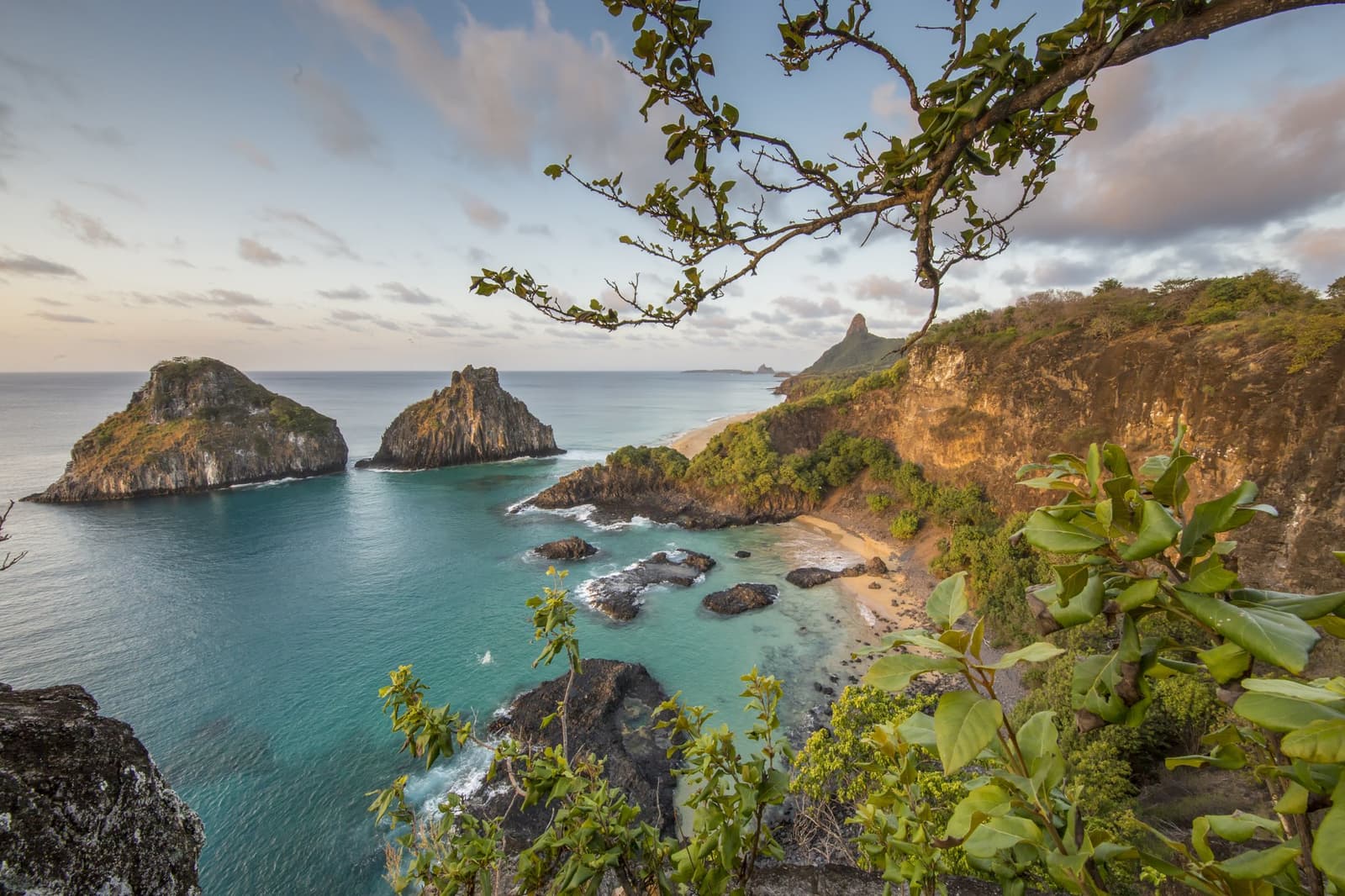 That moment when you realize the postcard didn't exaggerate. Aerial view of turquoise coves, golden cliffs and twin rock islets framing the coastline of Fernando de Noronha, the setting for a 2-Day Diving Package in Fernando de Noronha
