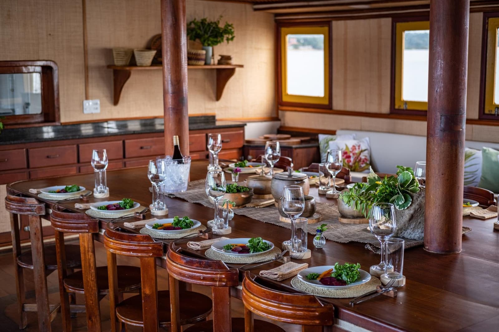 Good food, river views, and no dress code required. Elegantly set oval dining table aboard an Amazon cruise from Alter do Chão, with fresh salad plates, wine glasses, a bottle chilling on ice, rustic wooden stools, and river views through yellow-trimmed portholes