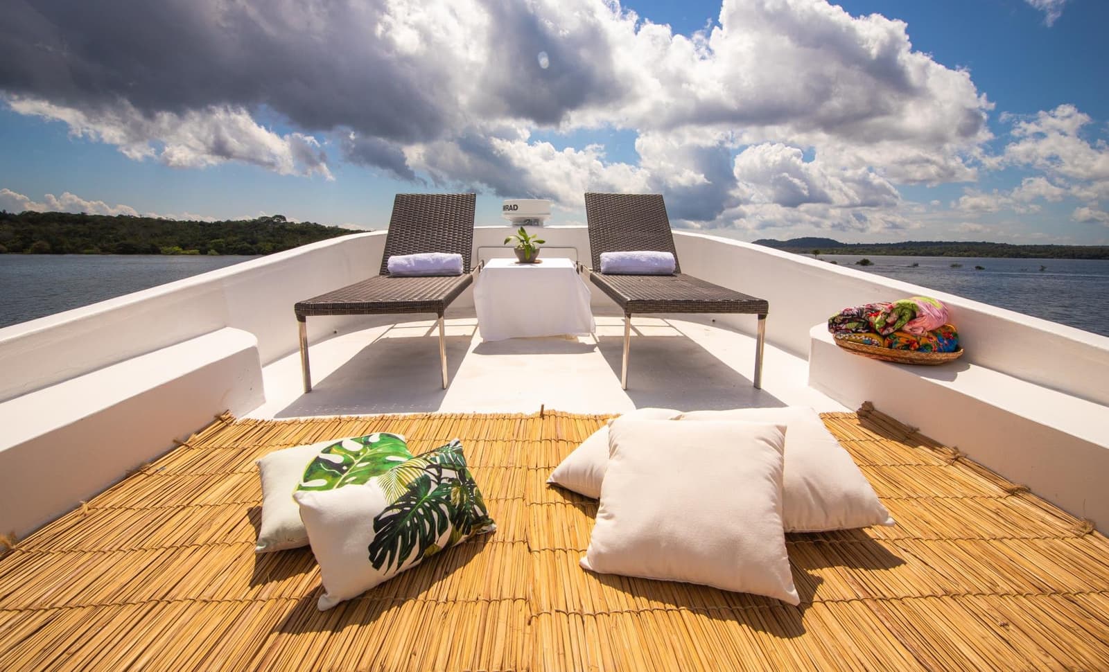 Your office for the afternoon, with better cloud cover than most. Two wicker sun loungers with rolled white towels on the open bow deck of an Amazon cruise from Alter do Chão, with tropical floor cushions, a bamboo mat, and forested riverbanks stretching under a dramatic cloudy sky