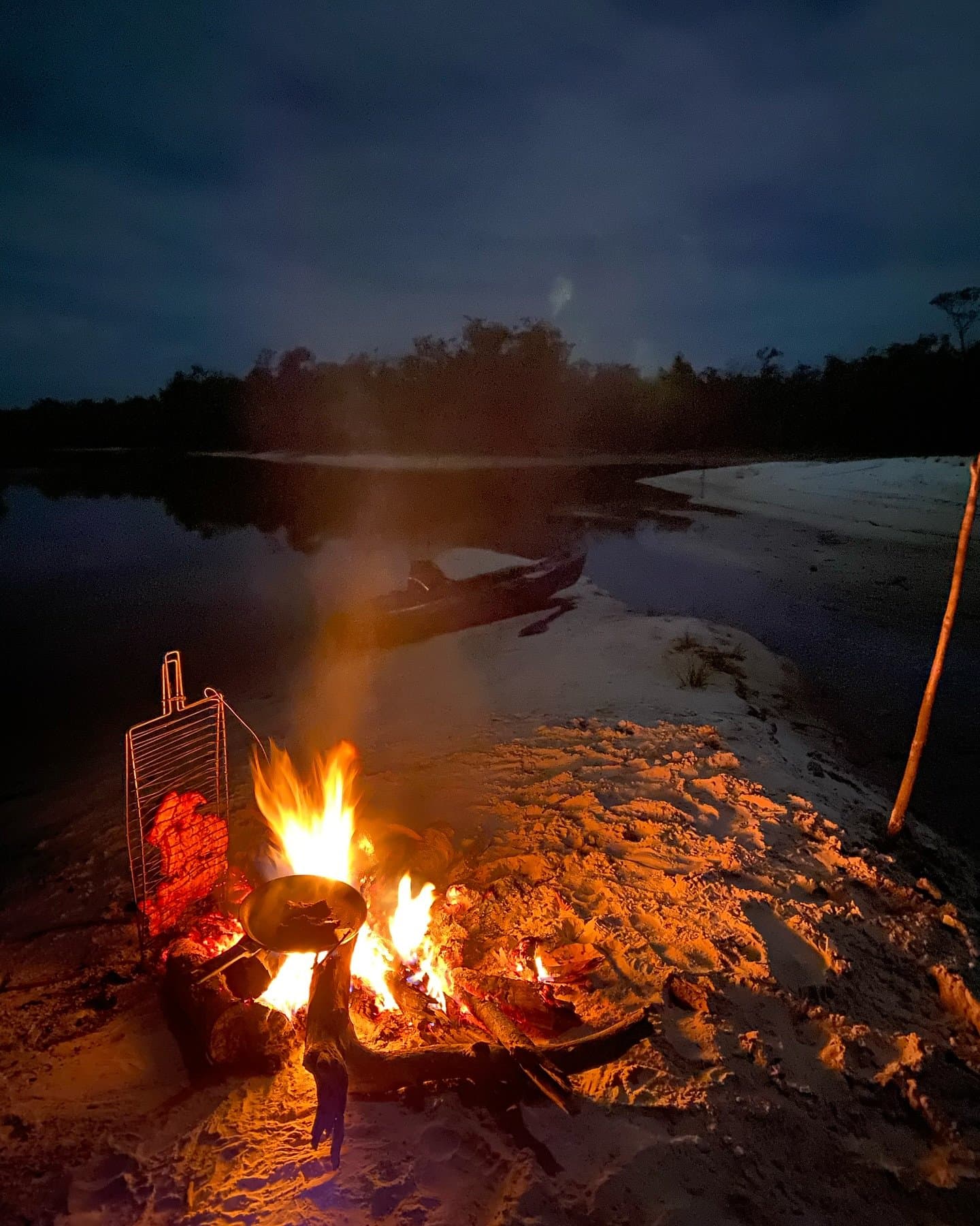 Dinner sorted, boat tied up, river doing its thing. A crackling campfire with a cooking pan and grill rack on a white sand riverbank at night, with a wooden canoe resting at the water's edge during an Amazon kayak tour in Brazil
