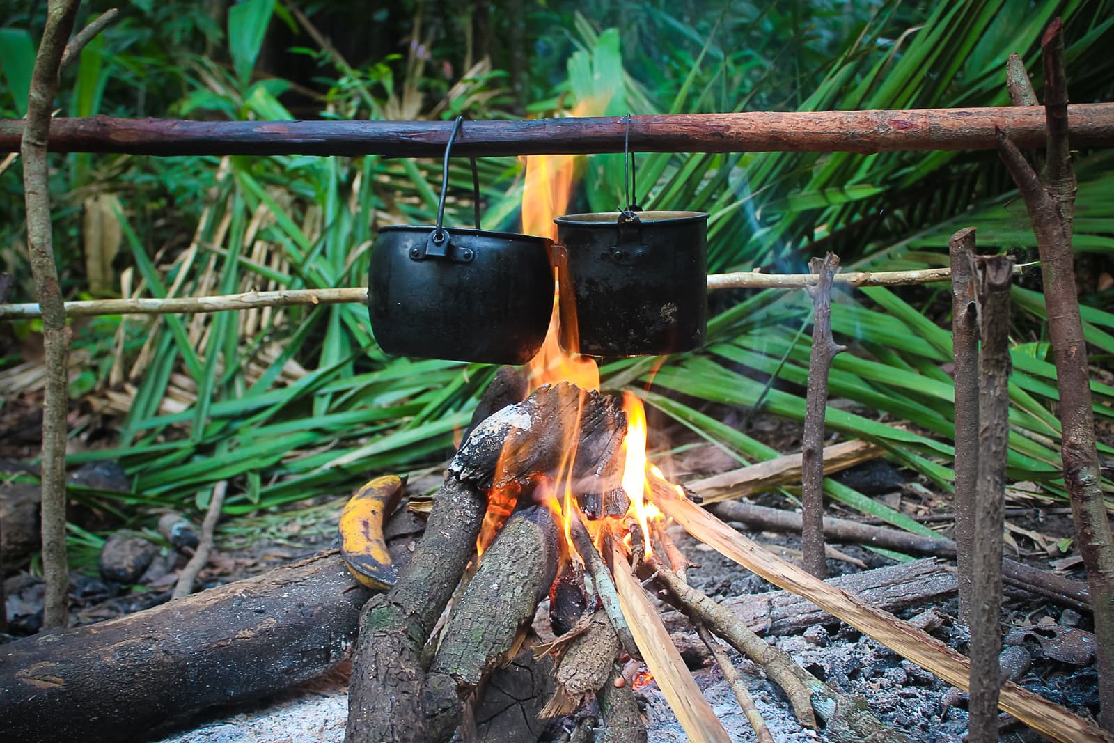 Dinner is on, and the jungle does not care about your schedule. Two blackened iron pots hanging over an open wood fire on a rustic stick frame during an Amazon trek and survival tour, surrounded by dense tropical vegetation