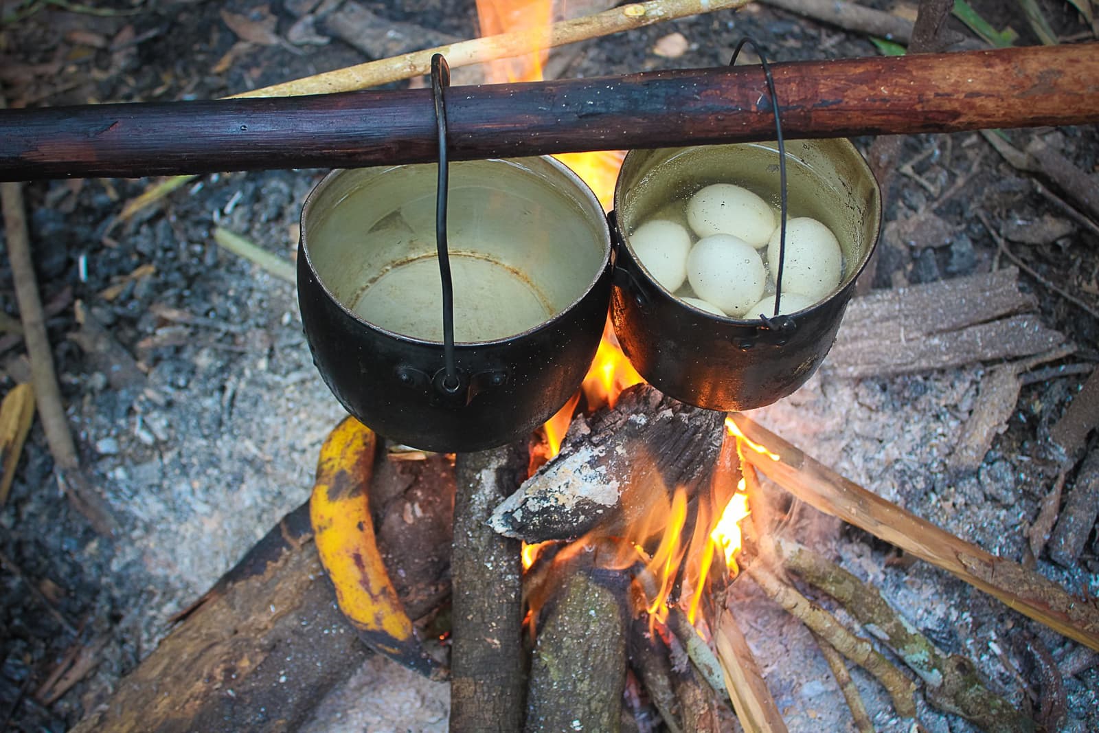 Dinner sorted, no kitchen required. Two blackened pots hanging over an open wood fire on a jungle survival Brazil camp, one holding eggs boiling in water