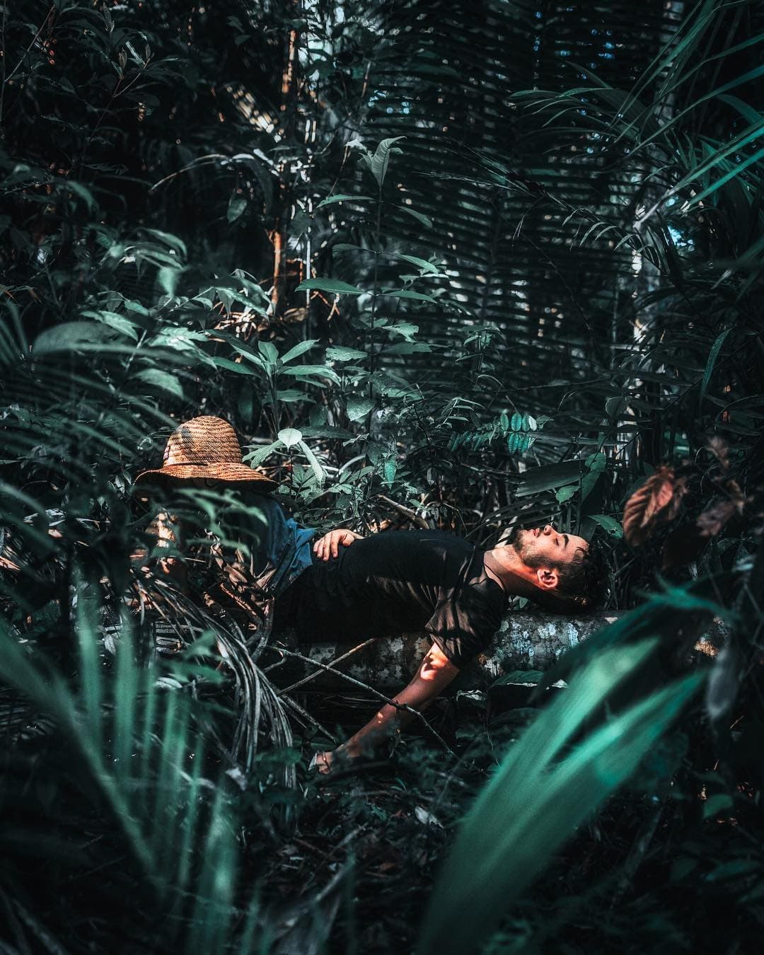 The forest floor has a way of insisting you slow down. A man lies back on a mossy log deep in dense rainforest undergrowth, his straw hat set aside, resting during an Amazon trek and survival tour