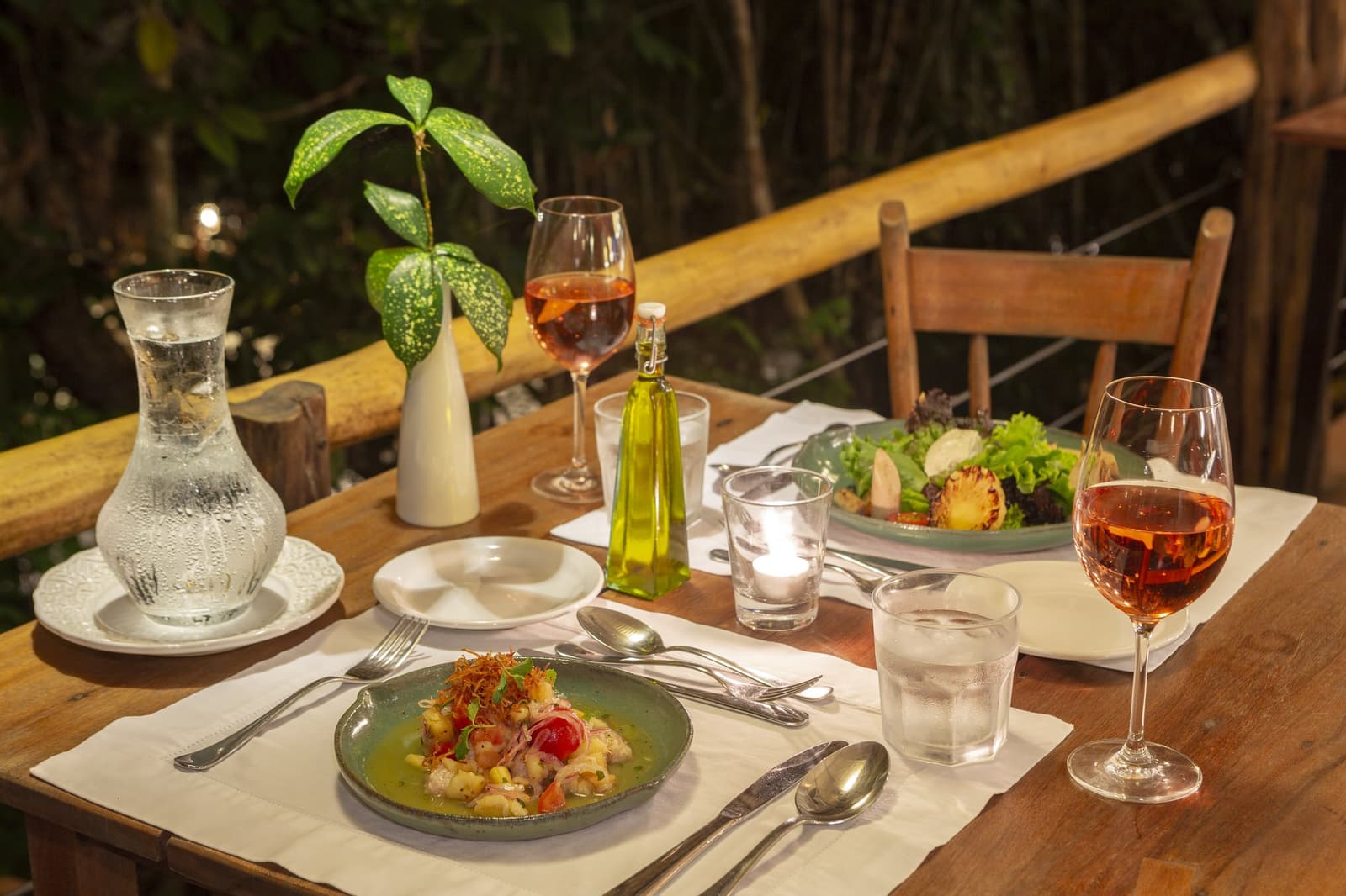 Good food, good wine, and the jungle just close enough to feel it. A candlelit outdoor dining table at Anavilhanas Jungle Lodge Brazil set with rosé wine, a green ceramic plate of seafood starter, fresh salad, olive oil and a tropical leaf centrepiece, overlooking the jungle on a bamboo-railed terrace