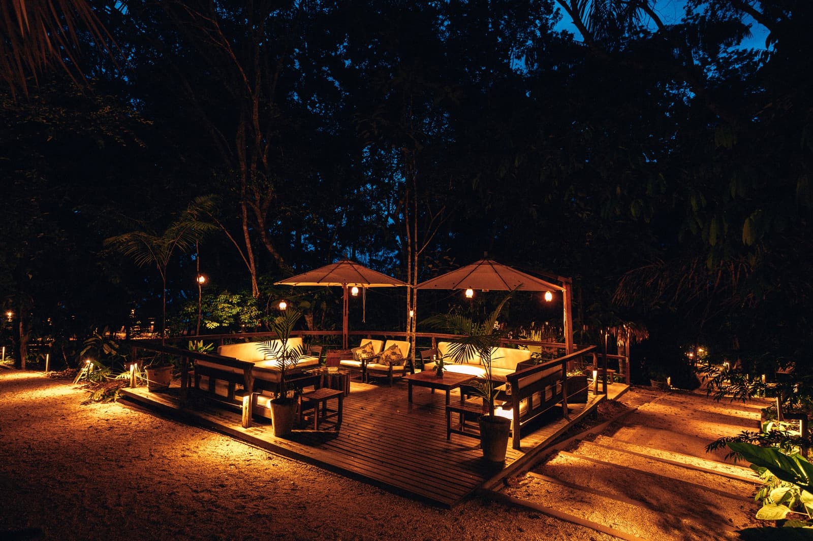 The kind of evening where the jungle does all the talking. An illuminated open-air deck at Anavilhanas Jungle Lodge Brazil, with cushioned wooden sofas arranged beneath canvas umbrellas and warm lantern light glowing against the surrounding rainforest at dusk
