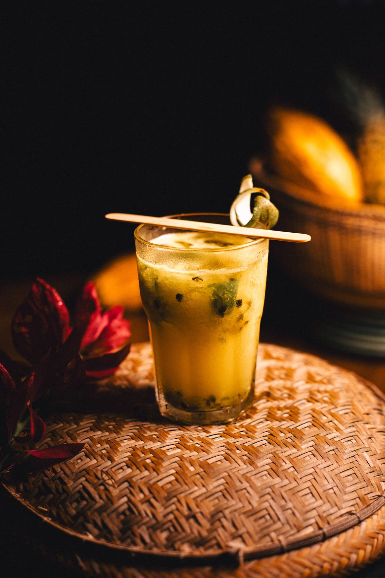 Passion fruit does all the talking here. No complaints. A golden passion fruit cocktail with visible seeds served in a glass on a woven rattan tray at Anavilhanas Jungle Lodge Brazil, garnished with a wooden stirrer and a curled tropical leaf, with a basket of yellow fruit softly lit in the background