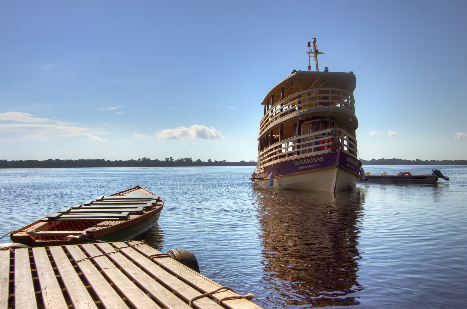 Your floating base camp, ready and waiting on the river. The Anavilhanas Jungle Lodge Brazil riverboat moored on the calm black waters of the Rio Negro, viewed from a wooden dock with a traditional wooden canoe in the foreground