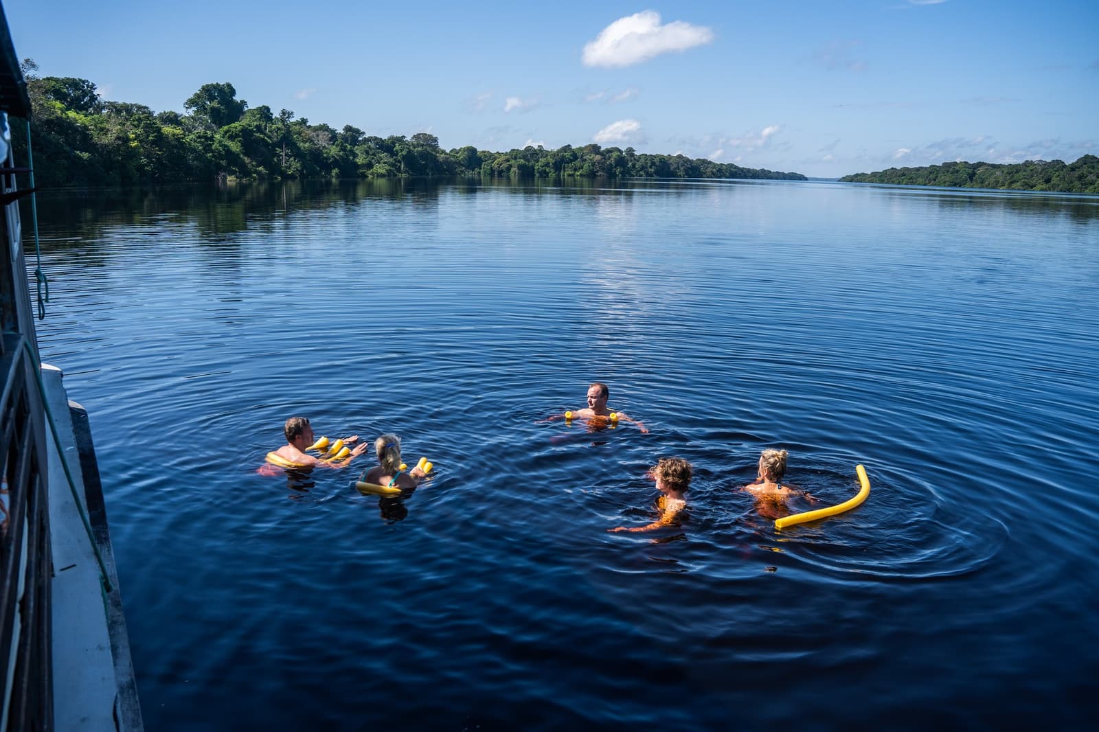 Yes, you really do swim in the Amazon. Yes, it is worth it. Five guests swimming in the dark waters of the Rio Negro beside a traditional boat at Anavilhanas Jungle Lodge Brazil, with yellow foam noodles and dense rainforest lining both riverbanks under a blue sky