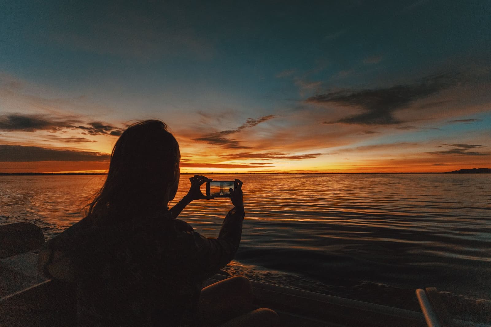 Some sunsets demand a second frame, one on the water, one on your phone. A traveller silhouetted against a blazing orange and teal sunset photographs the wide river from a boat at Anavilhanas Jungle Lodge Brazil