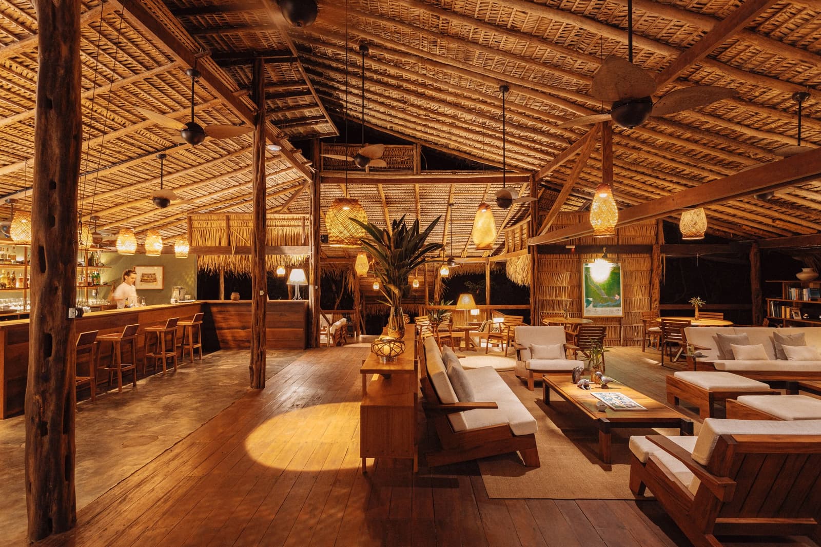 The kind of bar where you lose track of the hour, and don't mind one bit. Warmly lit open-air lounge at Anavilhanas Jungle Lodge Brazil, featuring a thatched palm roof, woven pendant lights, hardwood floors, linen sofas and a wooden bar with a bartender in the background