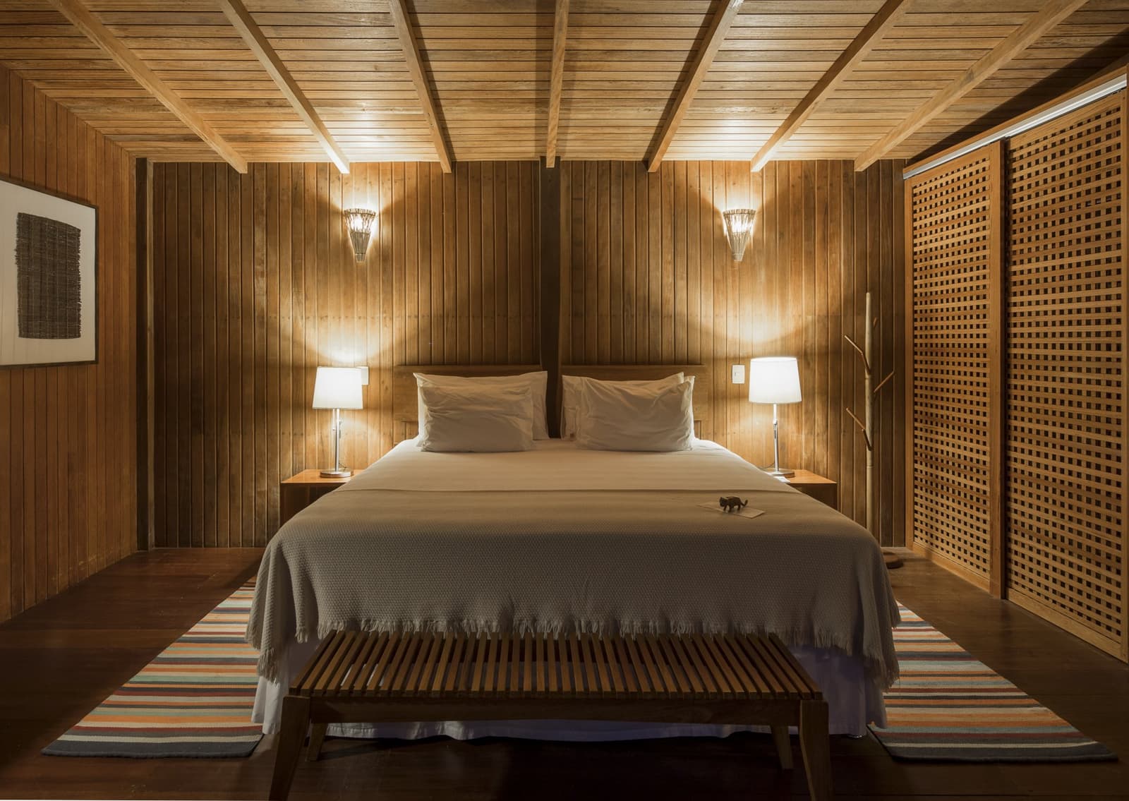 Warm wood everywhere, and the forest just outside the window. A warmly lit double bedroom at Anavilhanas Jungle Lodge Brazil, featuring floor-to-ceiling timber-panelled walls, a king bed with white linens and a grey fringed throw, twin bedside lamps, latticed wooden screens, and a striped rug on dark hardwood floors