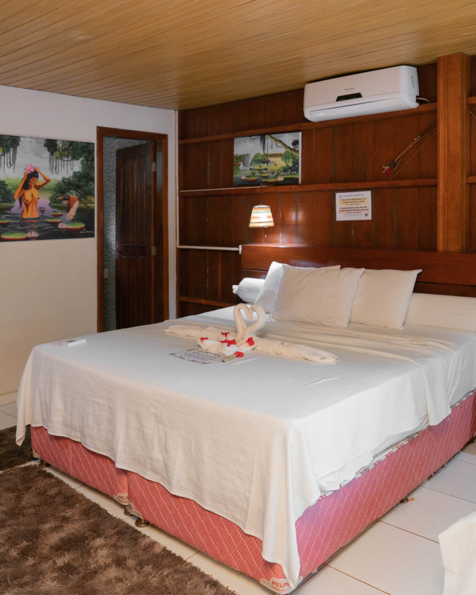 Jungle comfort: swan-folded towels, handpainted Amazon art, and a good night's sleep waiting. Photo: Yuri Panichi A double bed with white linens and towel-folded swans on the mattress, set in a cozy lodge room with a wooden headboard wall, warm lamp, Amazon-themed artwork, and air conditioning â comfortable accommodation included in the brazilian amazon jungle tour