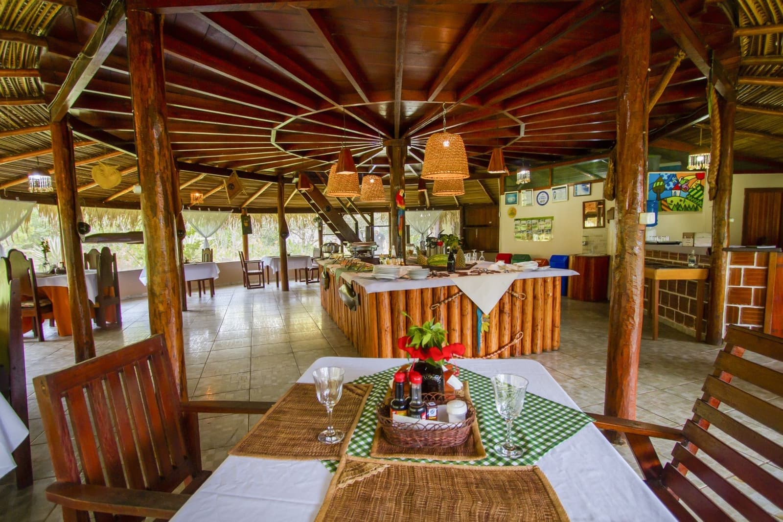 Rustic elegance under a thatched roof â meals served deep in the rainforest. Open-air thatched dining pavilion at an Amazon jungle lodge, with rustic log columns, wicker pendant lights, white-clothed tables, and a buffet counter set with plates and tropical fruit