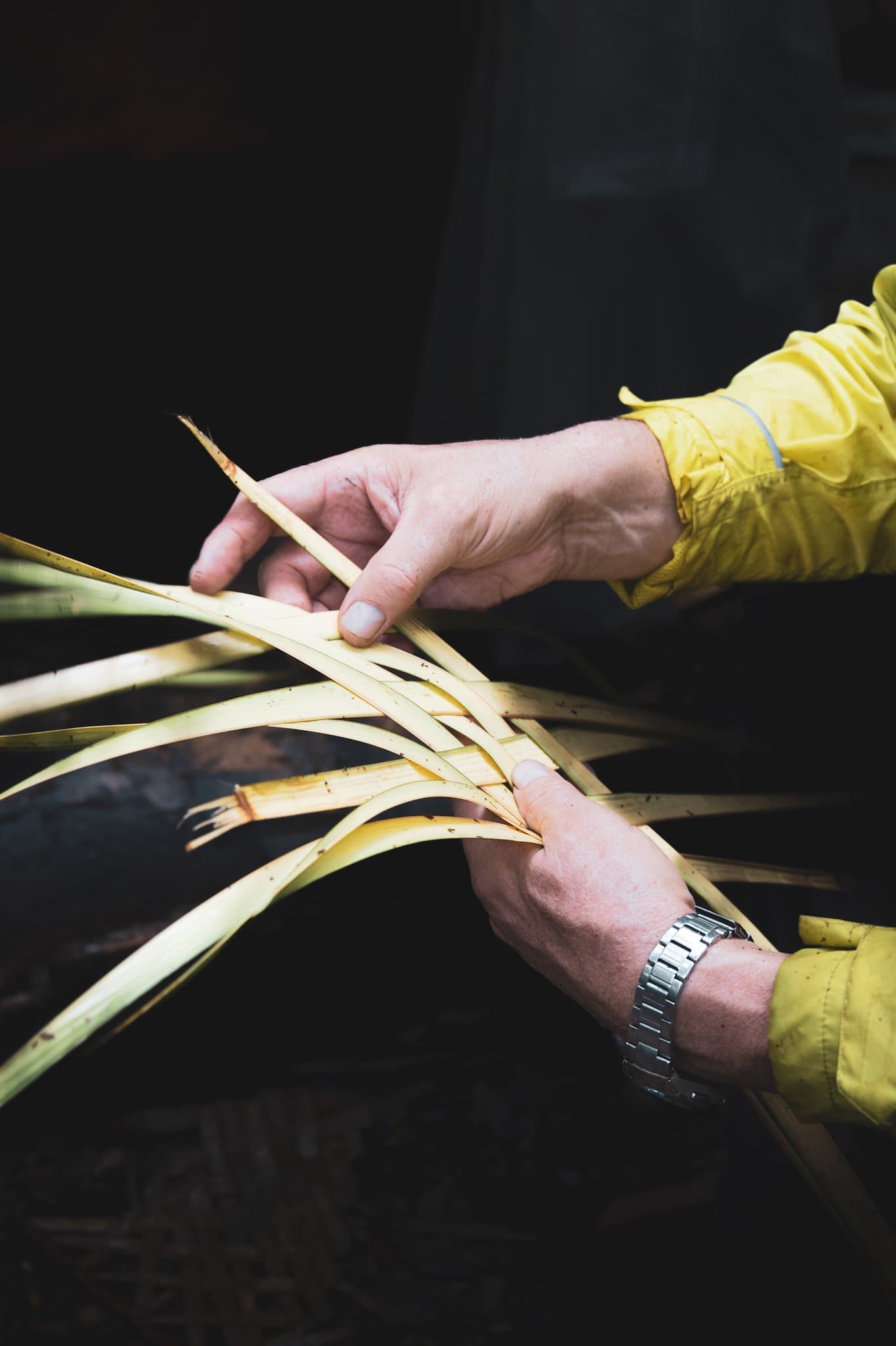 Nimble fingers, living fibers â jungle craft learned hands-on. Close-up of two hands in a yellow jacket weaving pale green palm leaf strips together during a brazilian amazon jungle tour survival skills lesson