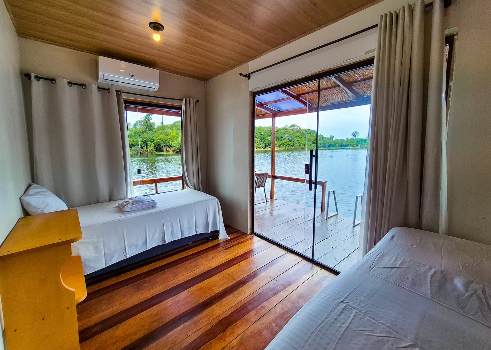 Sleep just metres from the river, forest pressing in on every side. Twin-bed lodge room with polished hardwood floors, air conditioning, and a sliding glass door opening onto a covered deck overlooking a calm river surrounded by dense rainforest â part of the brazilian amazon jungle tour accommodation