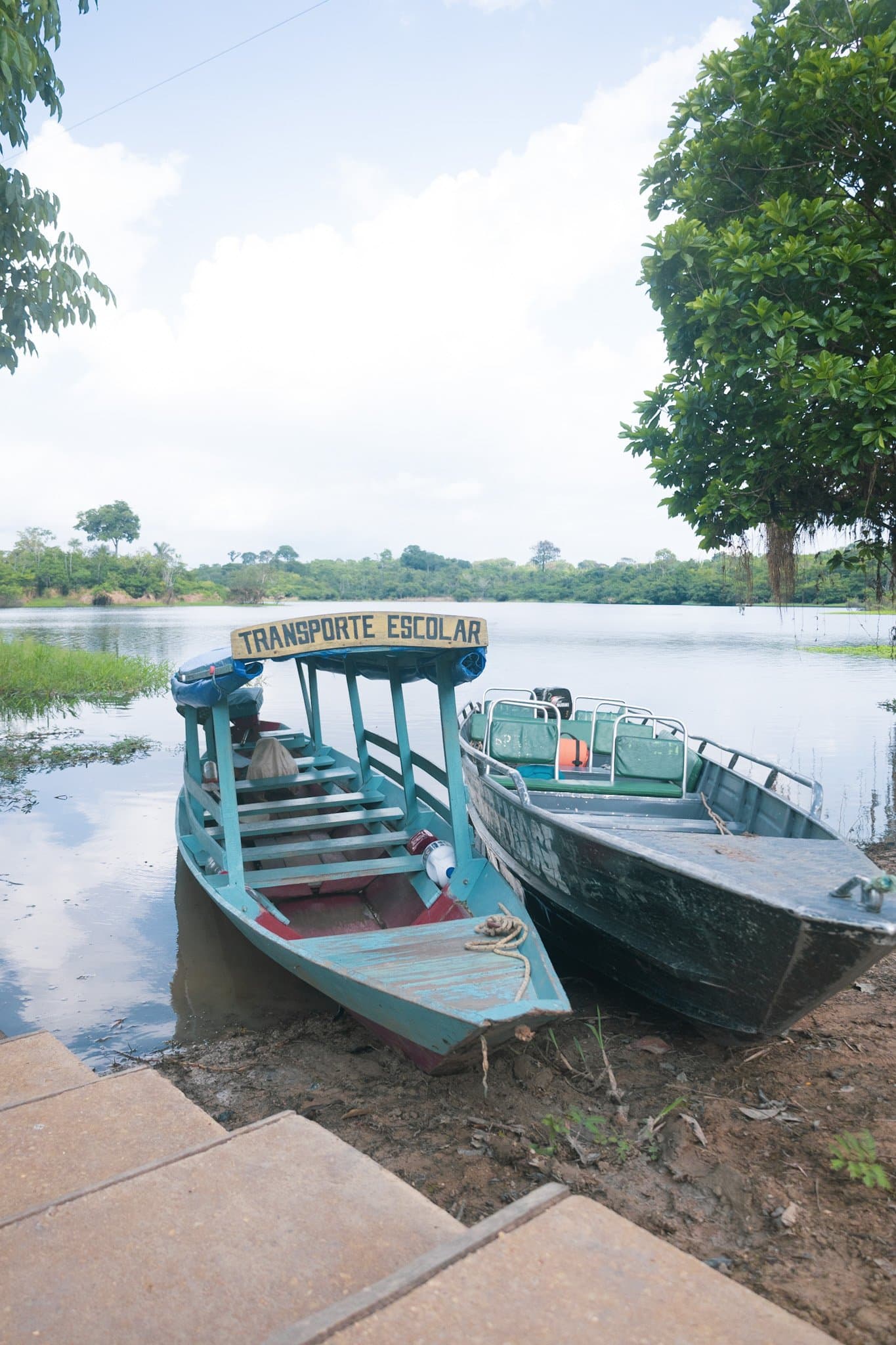 School boats on the river's edge â life in AmazĂŽnia runs on water. A turquoise wooden 'Transporte Escolar' boat and a second motorboat moored on a muddy riverbank, with a calm river and lush tropical vegetation stretching to the horizon â a scene from a brazilian amazon jungle tour
