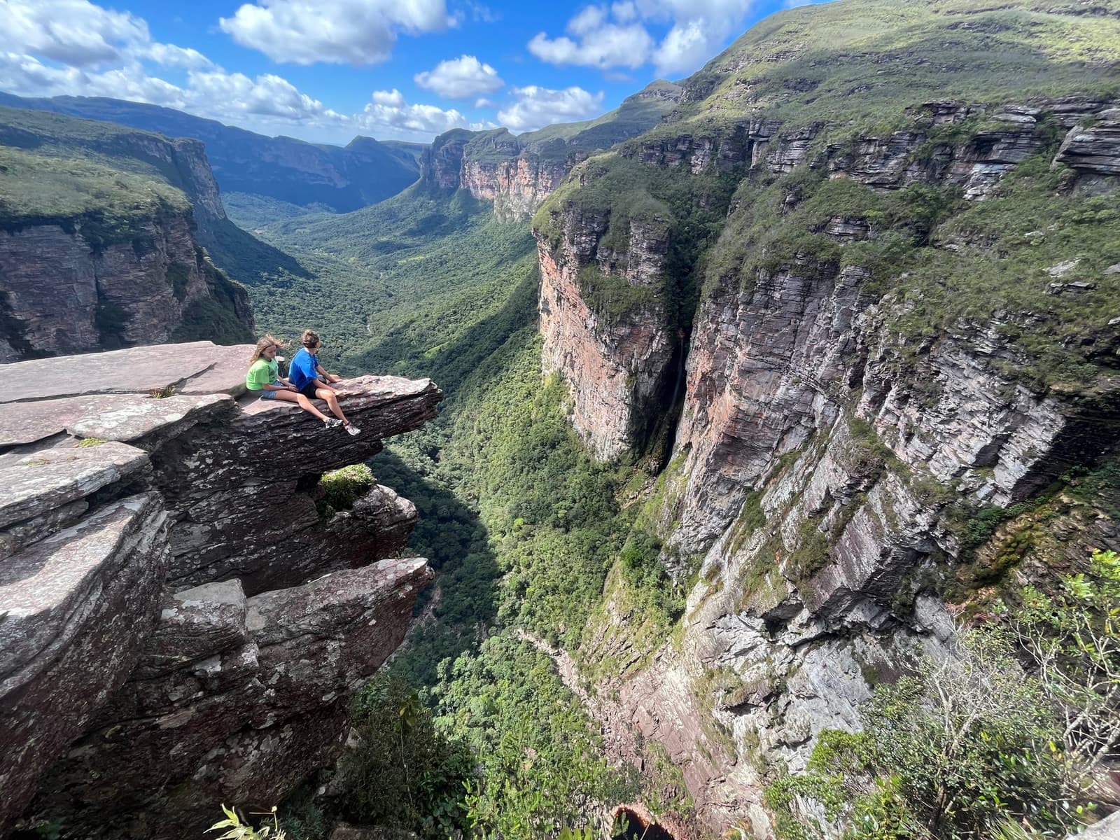 That ledge is absolutely real, and yes, their feet are dangling. Two hikers sitting on a jutting rock ledge above a deep canyon on a chapada diamantina hike, with layered sandstone cliffs and a forested valley stretching to the horizon