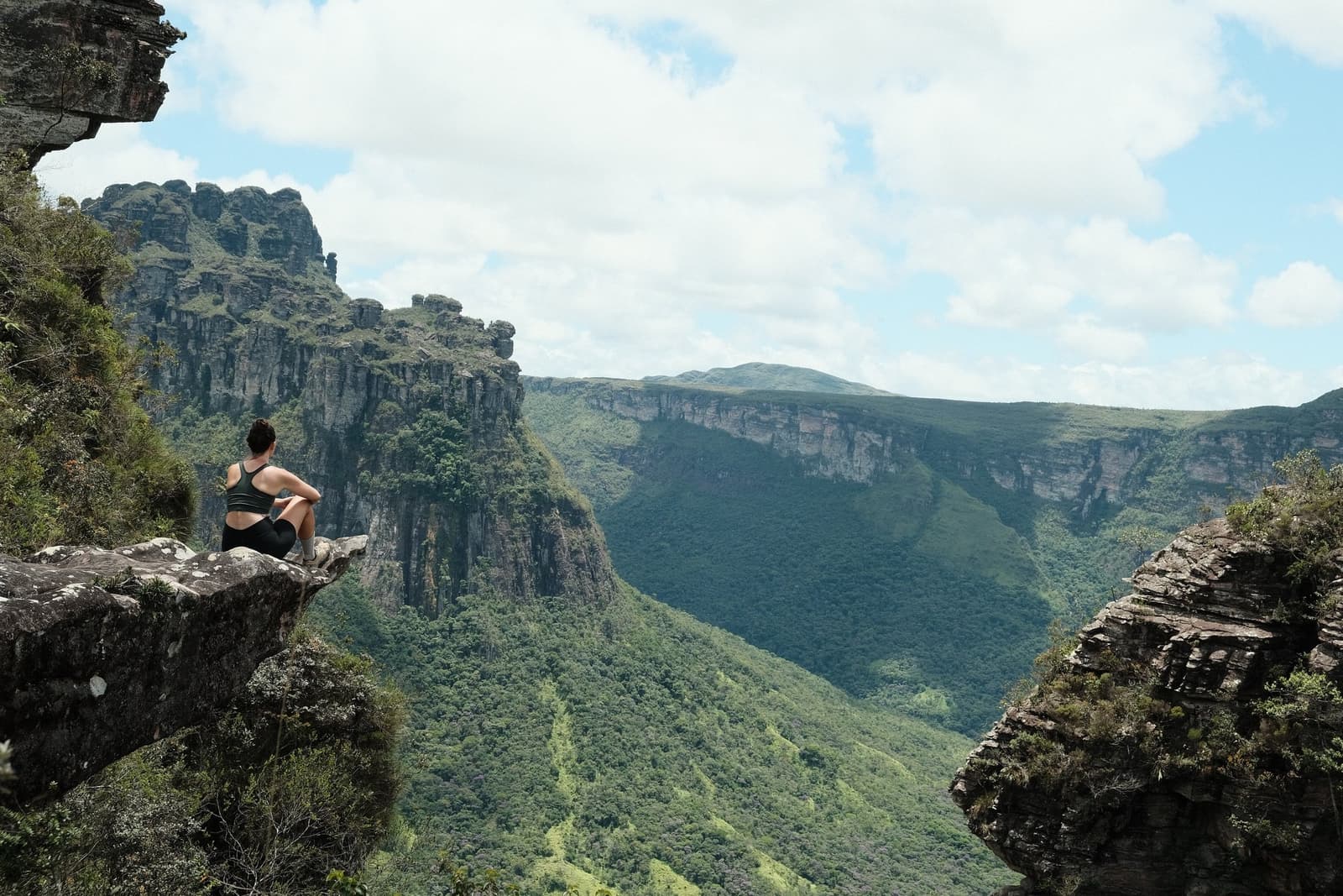 Some views earn their silence. This one takes a moment to sink in. A hiker sits on a rock ledge overlooking a vast green canyon with dramatic sandstone cliffs on a chapada diamantina hike
