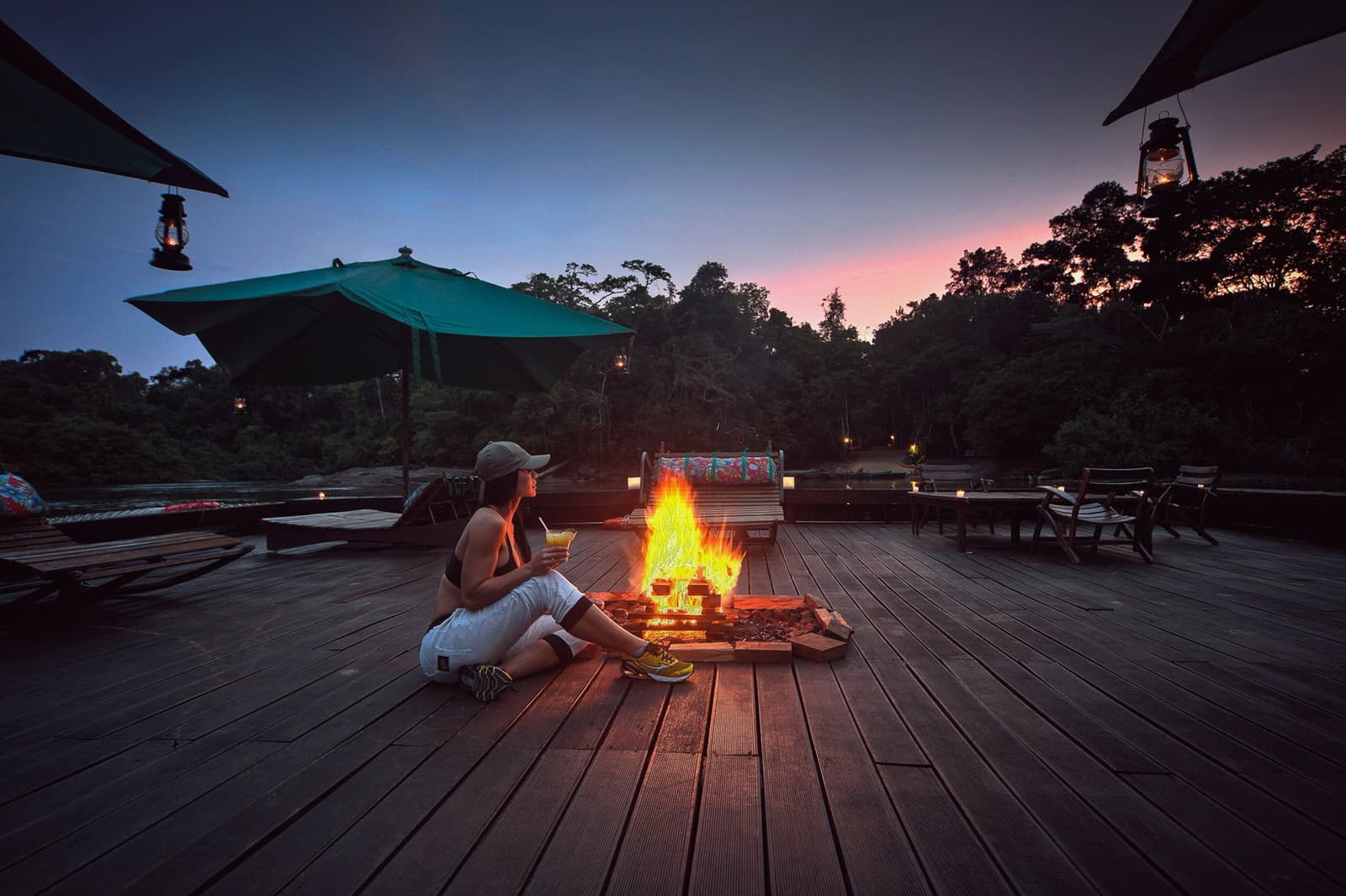 The Amazon night starts here, drink in hand, fire at your feet. A guest sits beside an open fire on the wooden deck at Cristalino Jungle Lodge, holding a cocktail as a pink-and-blue twilight sky glows above the rainforest canopy, with hanging lanterns lighting the scene