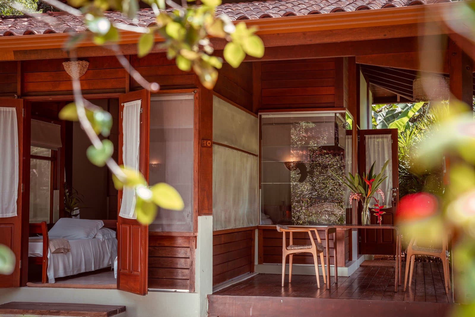 Your room opens straight into the forest. No lobby required. Timber bungalow at Cristalino Jungle Lodge with open doors revealing a white-linen bed, a covered veranda with wooden chairs, and tropical flowering plants framed by leafy branches