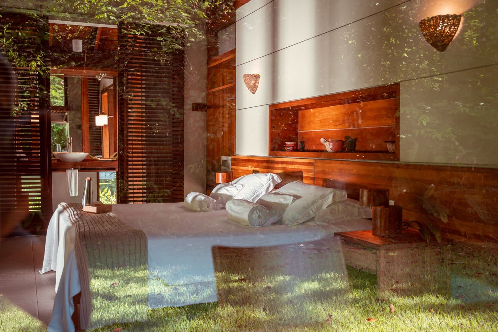 Sleep surrounded by the forest, sometimes almost literally. A Cristalino Jungle Lodge bungalow bedroom photographed through glass, reflecting surrounding greenery, with a double bed dressed in white linen, warm timber headboard with built-in shelving, woven wall sconces, and an en-suite vanity visible beyond wooden louvred shutters