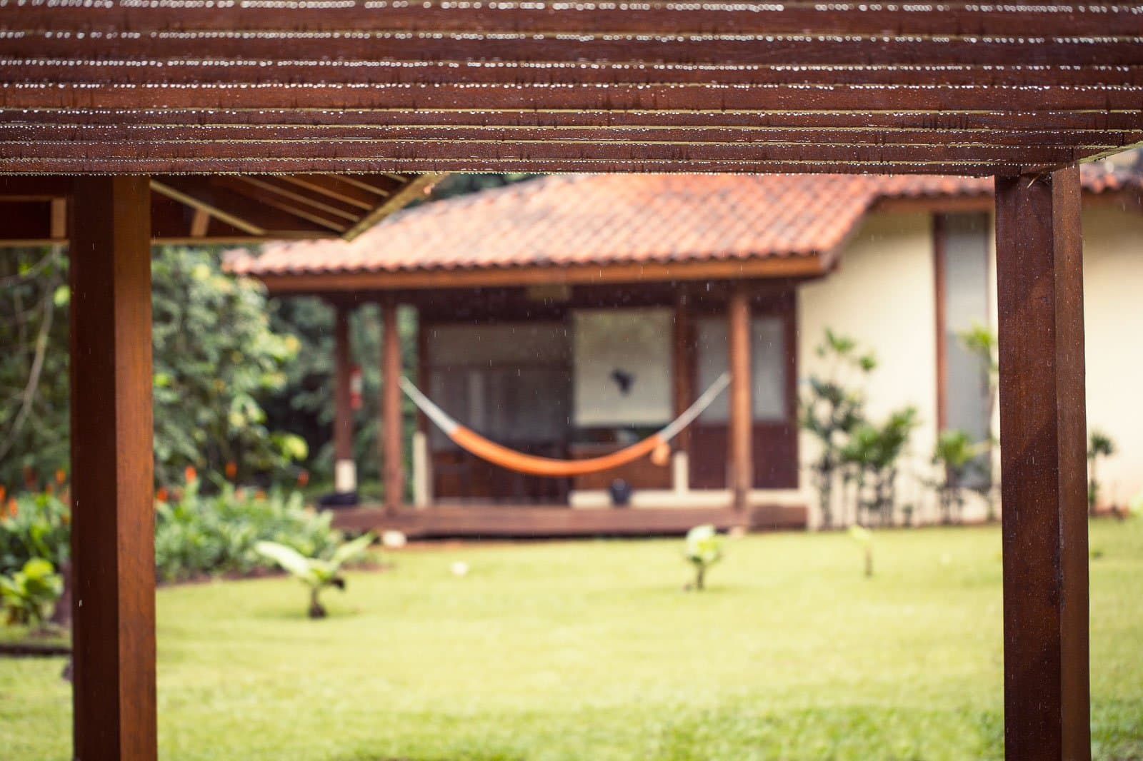 Rain on the roof, a hammock waiting. No better reason to slow down. Rain falling on a wooden pergola framing a view of a Cristalino Jungle Lodge bungalow with an orange hammock on its veranda and lush green lawns