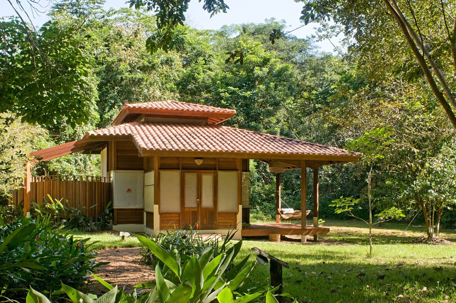 Your own patch of forest, with a hammock already waiting. A timber bungalow with a terracotta-tiled roof and a shaded wooden deck with a hammock, set among lush tropical forest at Cristalino Jungle Lodge
