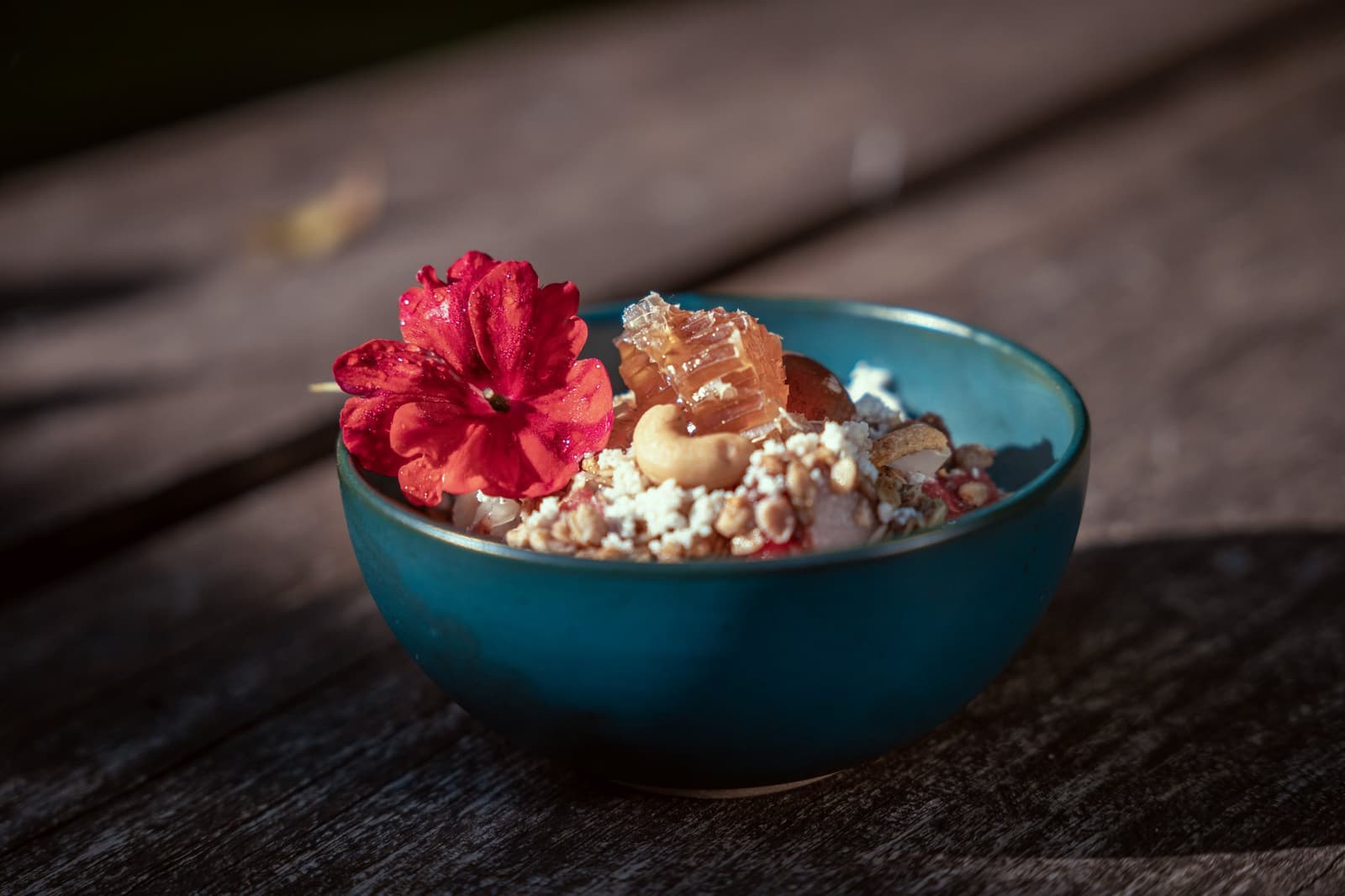 Breakfast in the Amazon comes with a flower and no apologies. A teal ceramic bowl at Cristalino Jungle Lodge filled with granola, nuts, honeycomb, and fresh fruit, garnished with a vivid red edible flower, served on a weathered wooden surface