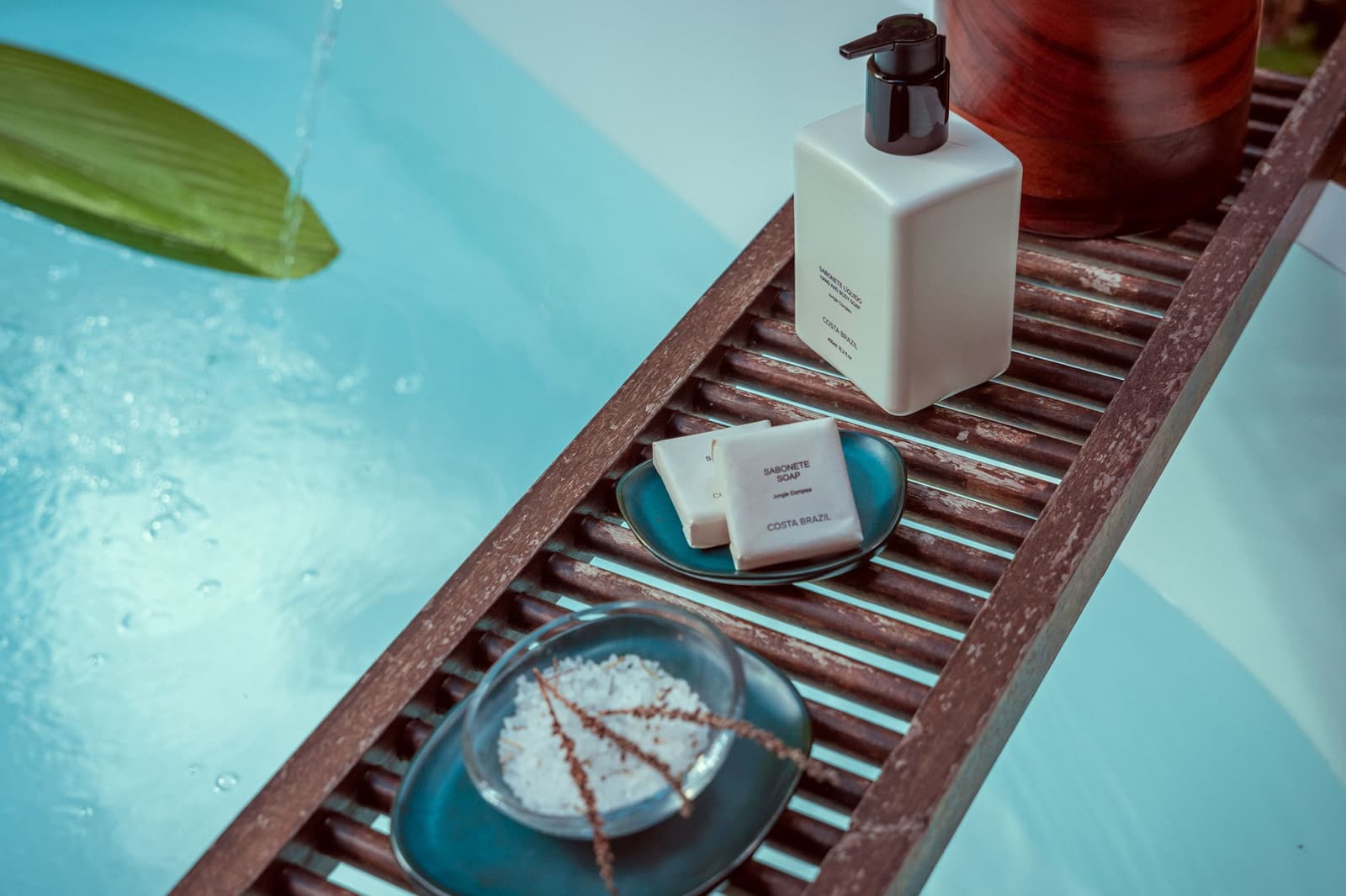 Little luxuries that hit differently after a morning in the forest. A wooden bath tray beside a turquoise pool holds Costa Brazil soap bars, a pump dispenser, and a dish of bath salts with a dried leaf at Cristalino Jungle Lodge