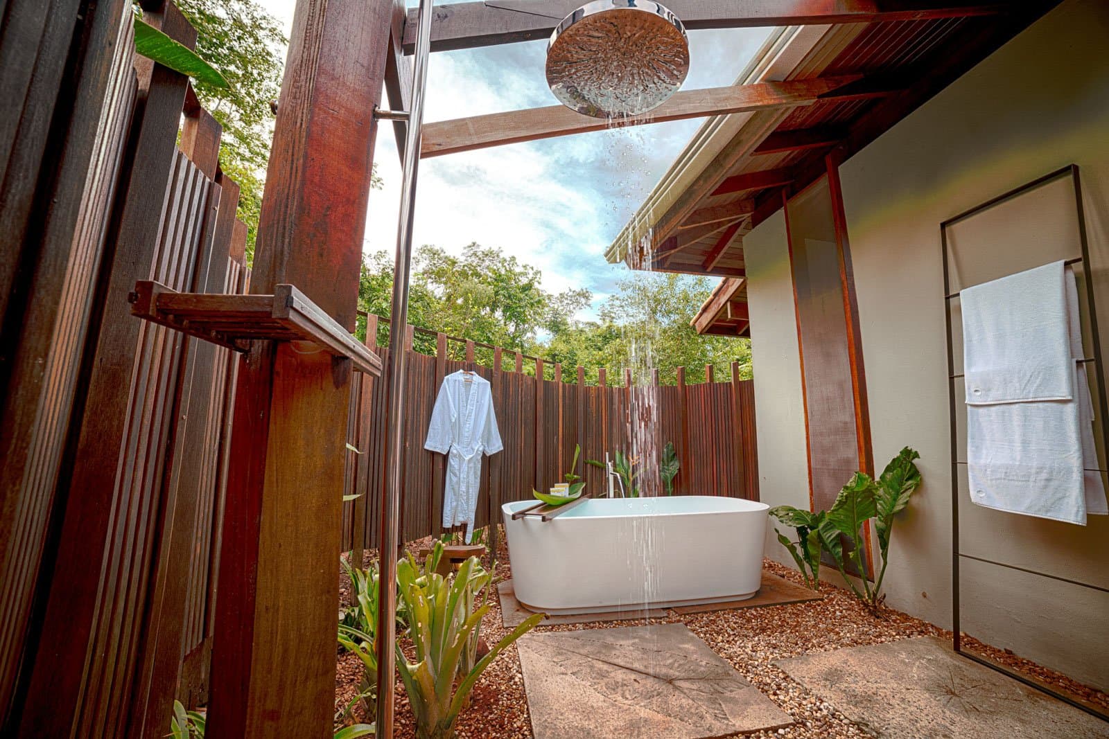 A hot bath under the Amazon sky, some comforts are non-negotiable. An open-air bathroom at Cristalino Jungle Lodge featuring a white freestanding soaking tub beneath a cascading rainfall showerhead, surrounded by tropical plants and warm hardwood screens with a forest canopy visible overhead