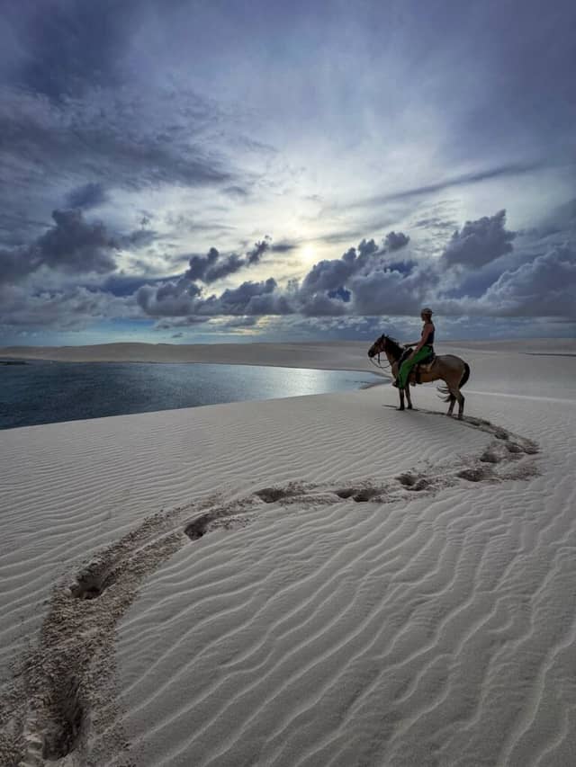 A solo rider crosses the white sand dunes of Lençóis Maranhenses National Park on horseback, with a rainwater lagoon stretching behind her. A solo rider crosses the white sand dunes of Lençóis Maranhenses National Park on horseback, with a rainwater lagoon stretching behind her.