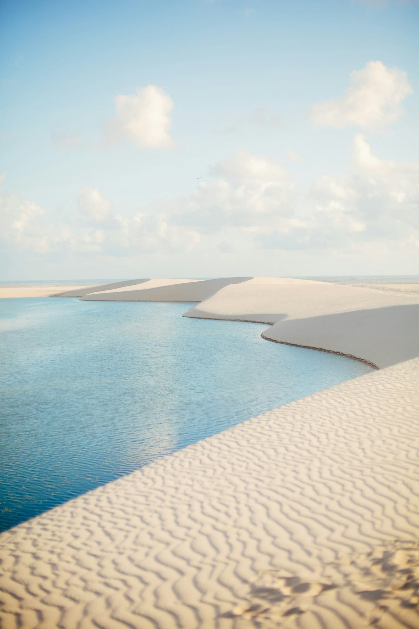 Sand and water taking turns deciding who's in charge. Turquoise rainwater lagoon nestled between sweeping white sand dunes under a soft blue sky in Lençóis Maranhenses, a highlight of the horseback riding Lençóis Maranhenses tour