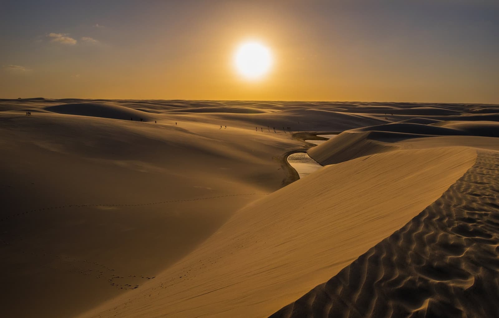 Sand, water, and a sun that takes its time going down. Golden sunset over the vast white sand dunes of Lençóis Maranhenses, with a small lagoon winding between dune ridges and tiny silhouettes of visitors visible in the distance on this horseback riding Lençóis Maranhenses tour landscape