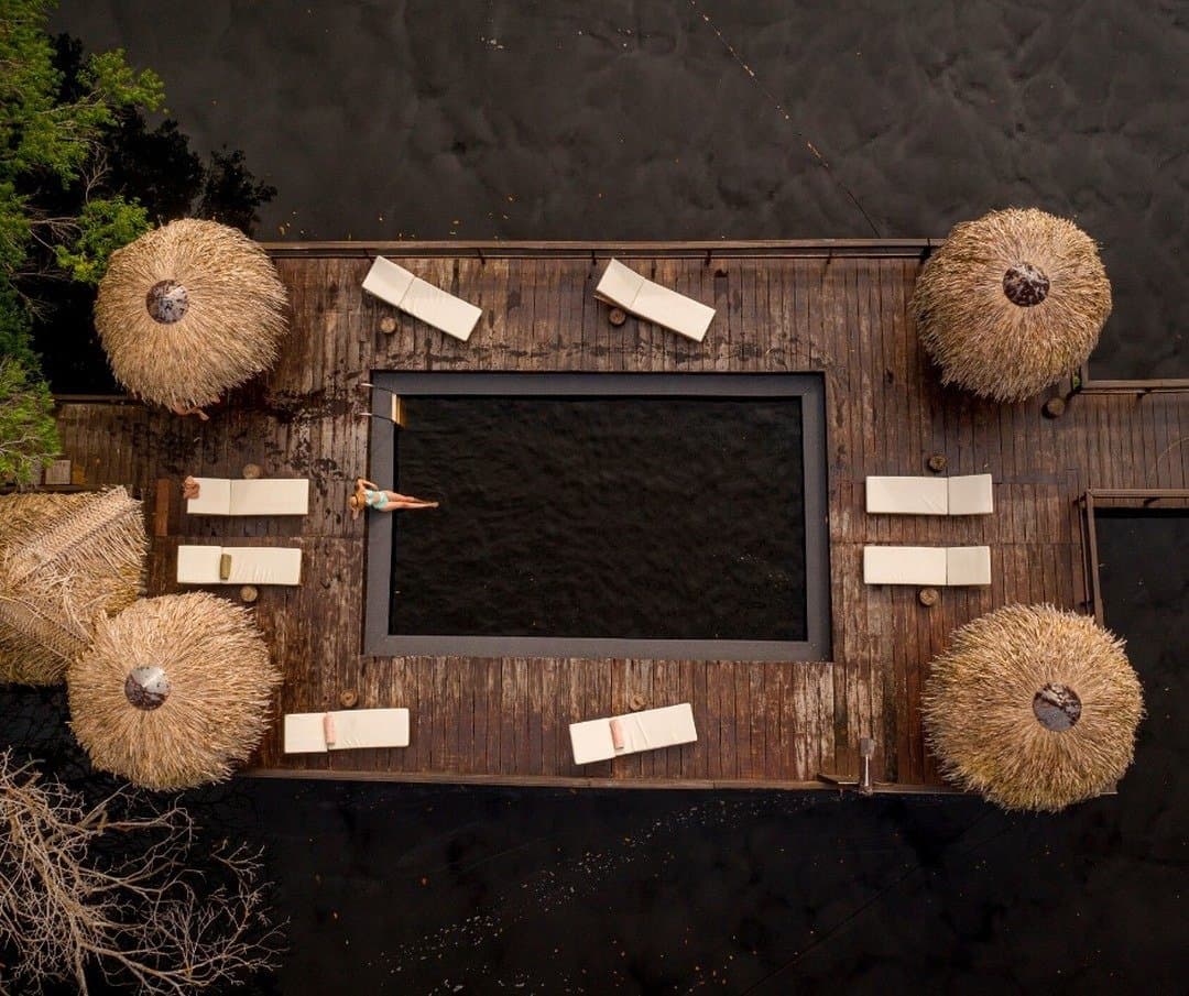 A pool open to the Amazon river, no chlorine, no walls, no problem. Aerial view of the Juma Amazon Jungle Lodge floating timber deck with a rectangular river pool, cream sun loungers, and thatched palm umbrellas surrounded by dark blackwater