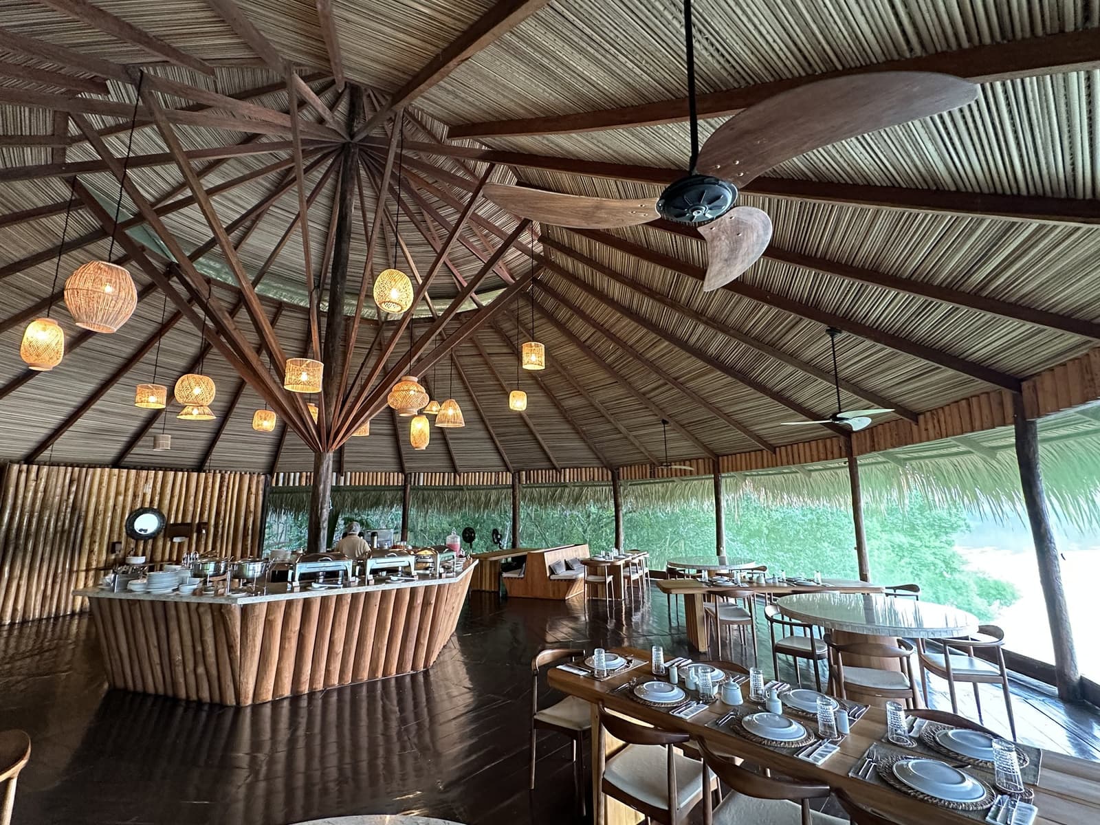 Dinner with the forest right at the edge of your table. The open-air restaurant at Juma Amazon Jungle Lodge, featuring a soaring thatched roof with radial timber beams, woven rattan pendant lights, a bamboo-clad buffet station, and wooden dining tables set with white crockery, open to lush green forest on all sides