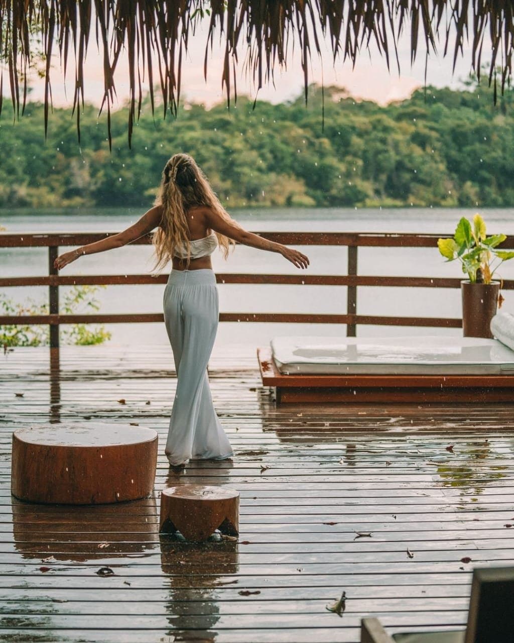Rain on a warm deck, arms open wide. Some moments need no plan. A woman in white stands arms outstretched on a rain-soaked deck at Juma Amazon Jungle Lodge, facing a calm river lined with dense green rainforest canopy under a thatched roof