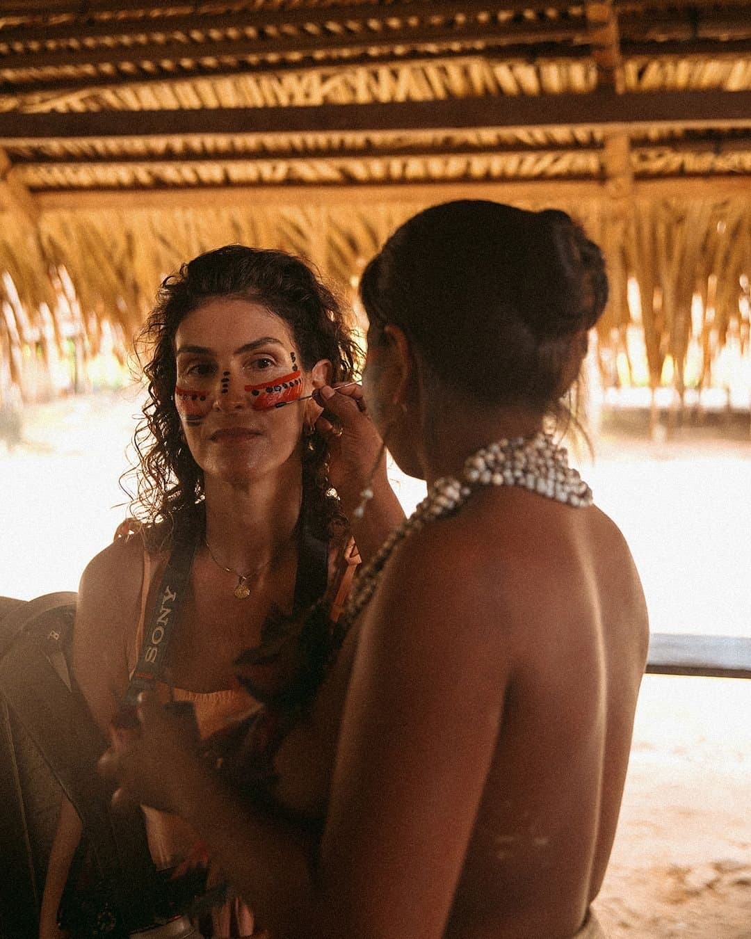A quiet moment of trust, painted on carefully. An indigenous woman applies traditional red and black face paint to a smiling traveller inside a thatched-roof open-air hut at Juma Amazon Jungle Lodge