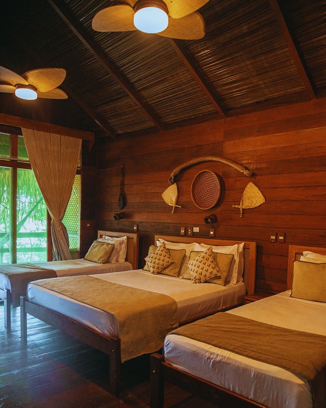 Rustic craft on the walls, the forest just outside the window, comfort that earns its setting. Triple bedroom at Juma Amazon Jungle Lodge with three wooden-framed beds, handwoven wall decor, a thatched ceiling with ceiling fans, and lush rainforest visible through the open balcony door