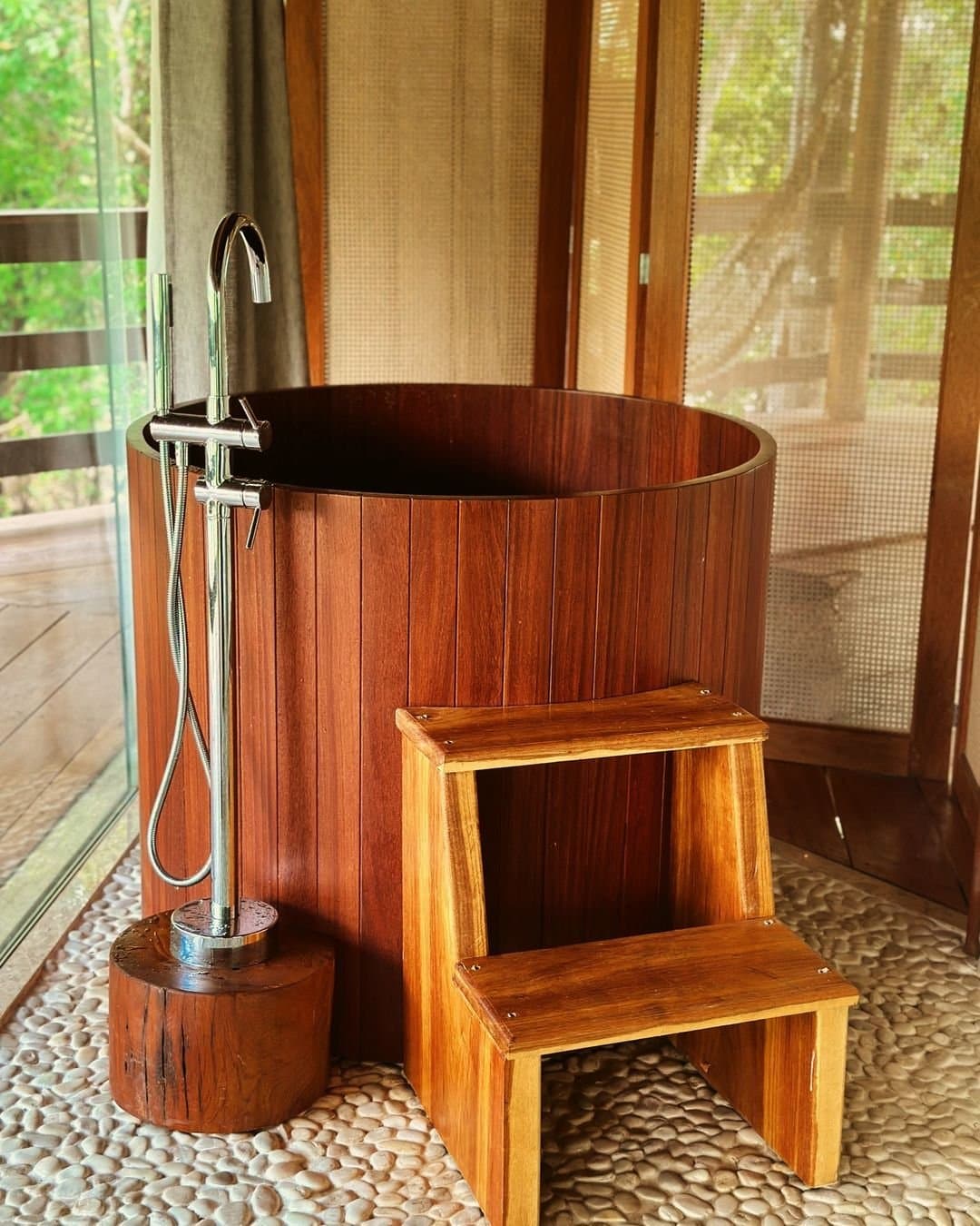 A soak in the forest, literally, the trees are right outside the window. A round polished-wood soaking tub with a chrome freestanding faucet and wooden step stool, set on a pebble-stone floor with forest-view windows at Juma Amazon Jungle Lodge