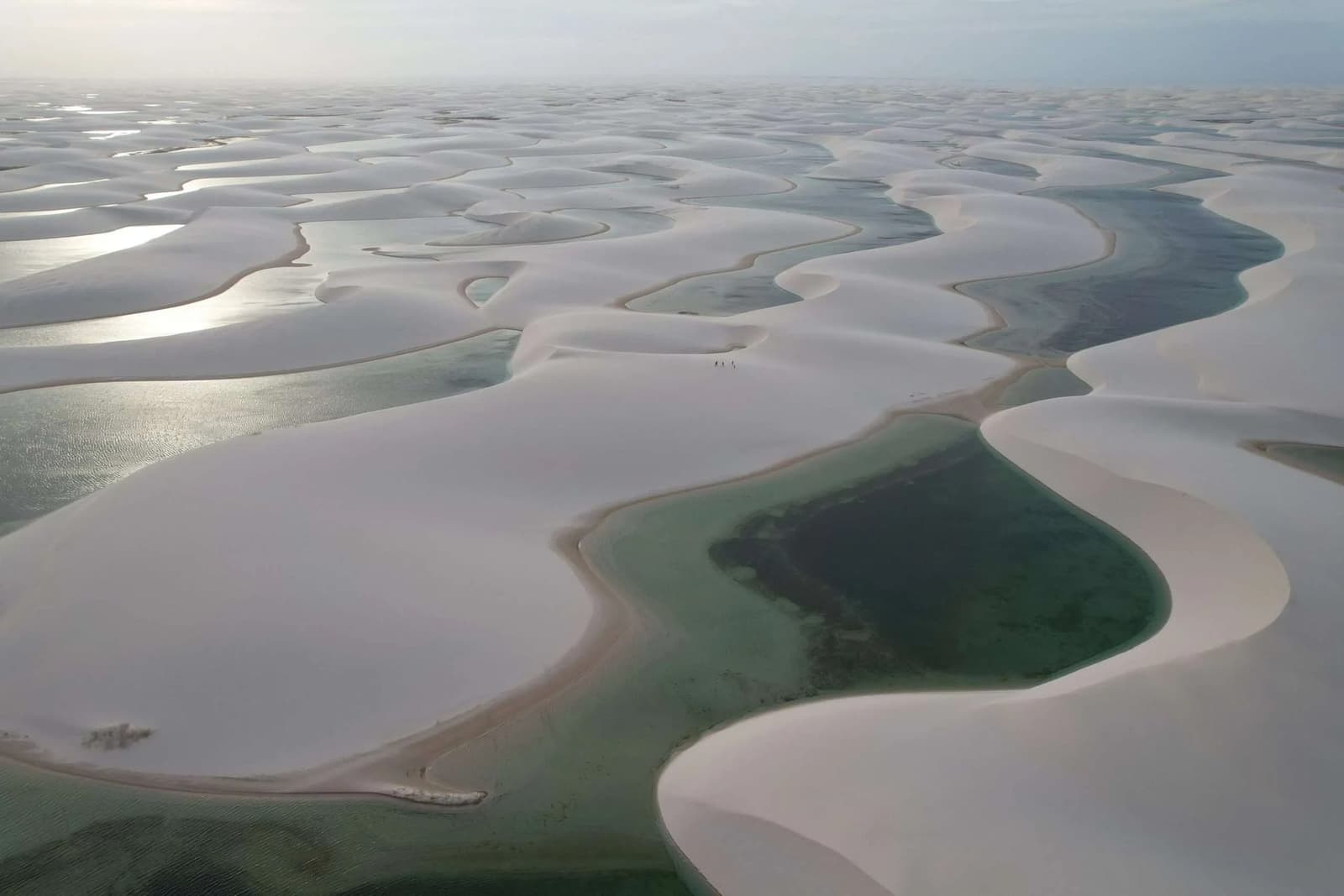 Sand dunes with their own lakes. Brazil never runs out of surprises. Aerial view of the Lençóis Maranhenses trip landscape, showing endless white sand dunes interspersed with dark-green and turquoise freshwater lagoons stretching to the horizon, with tiny human figures visible in the middle distance