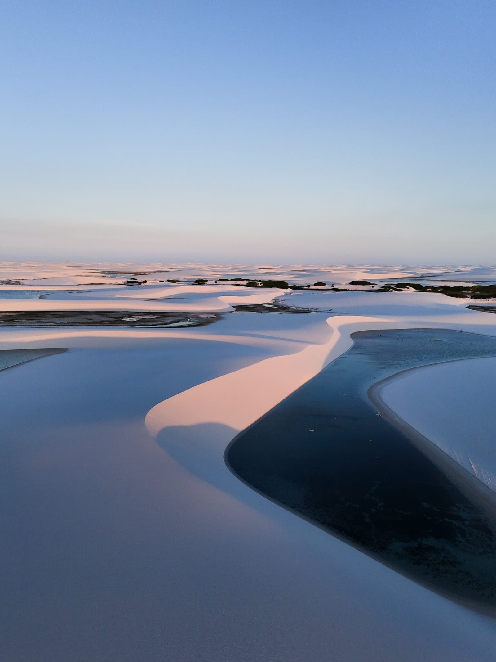 Pink light, white sand, cold water, the Lençóis at their most quietly dramatic. Aerial view of white sand dunes and dark blue lagoons glowing pink under a dusky sky on a Lençóis Maranhenses trip