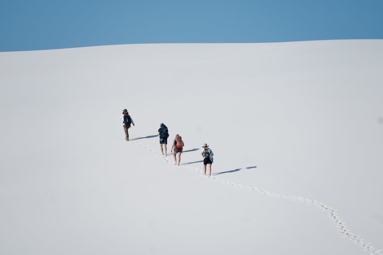 One foot in front of the other, the dunes have a way of making you forget time. Four backpackers climbing a vast white sand dune single-file on a Lençóis Maranhenses trip, leaving a trail of barefoot footprints under a clear blue sky